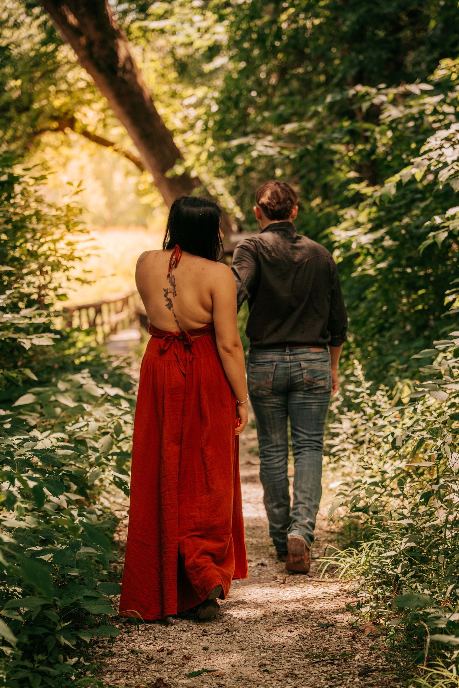 Couple walking on a forest path, woman in red backless dress with tattoo, man in jeans and gray shirt.