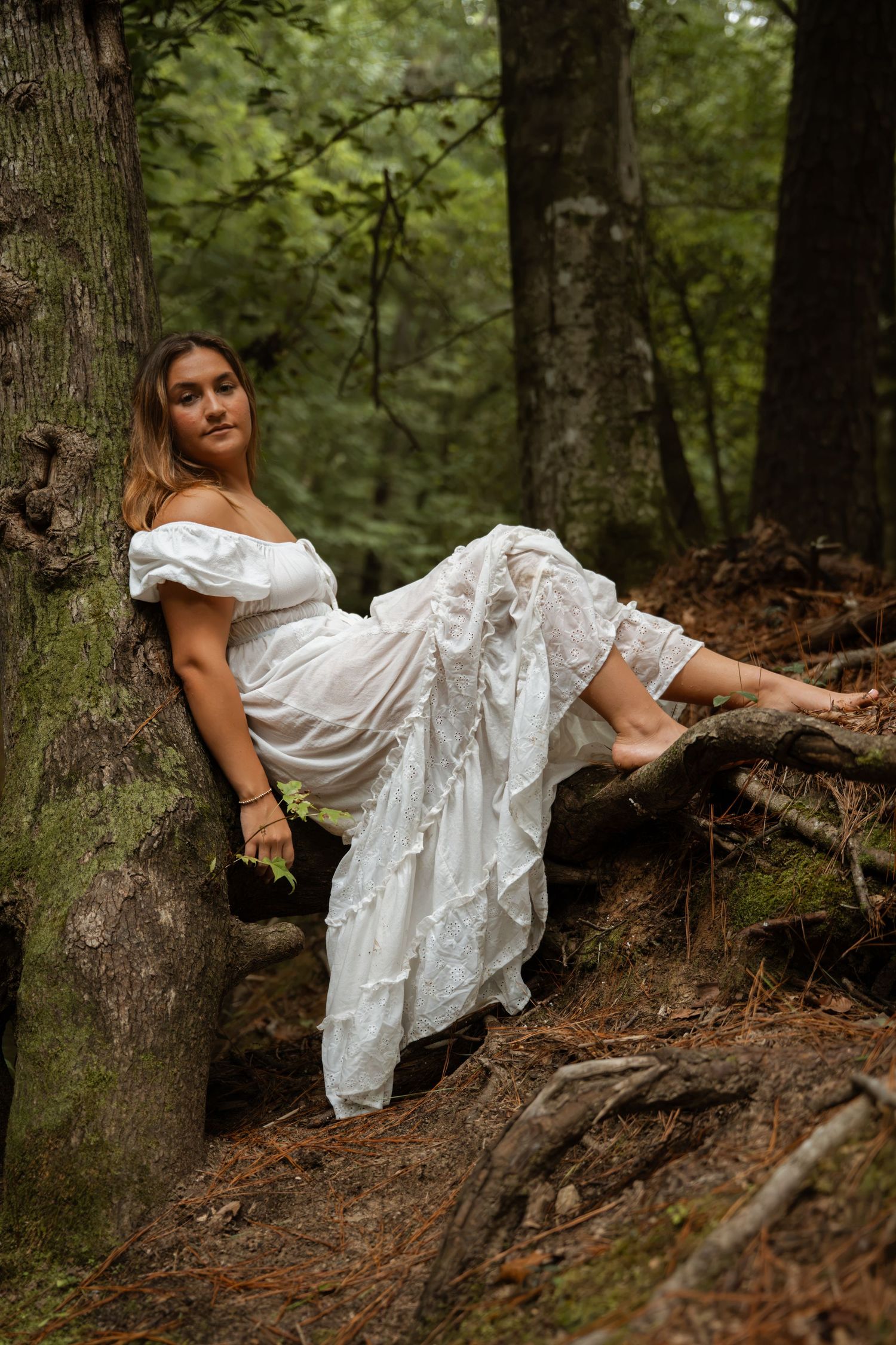 Woman in white off-shoulder dress resting against tree roots in a lush green forest setting.