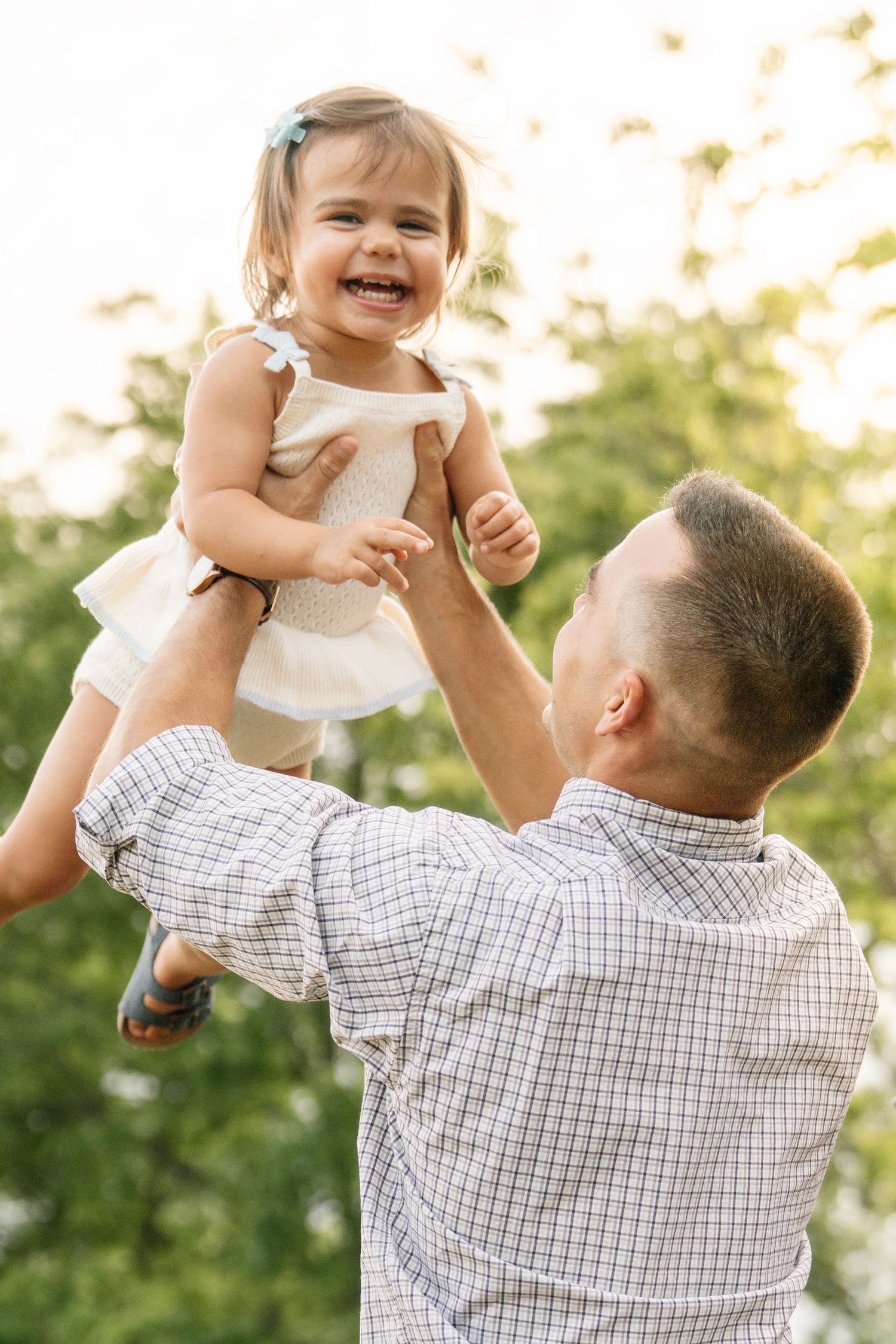 A joyful father lifts his laughing toddler daughter in a white dress outdoors in a sunny green park.