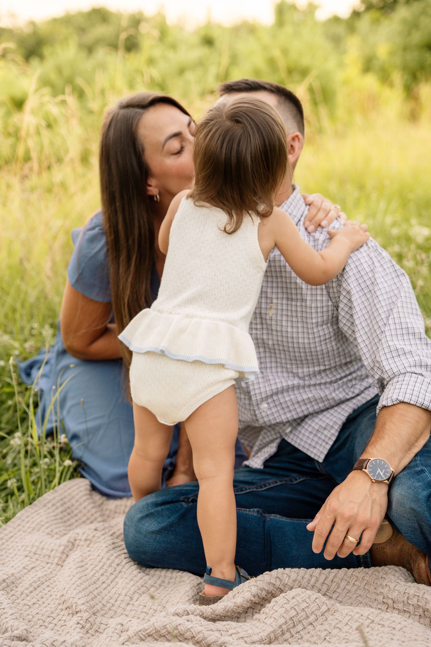 Mother kissing toddler daughter on cheek while sitting with father on blanket in grassy field.