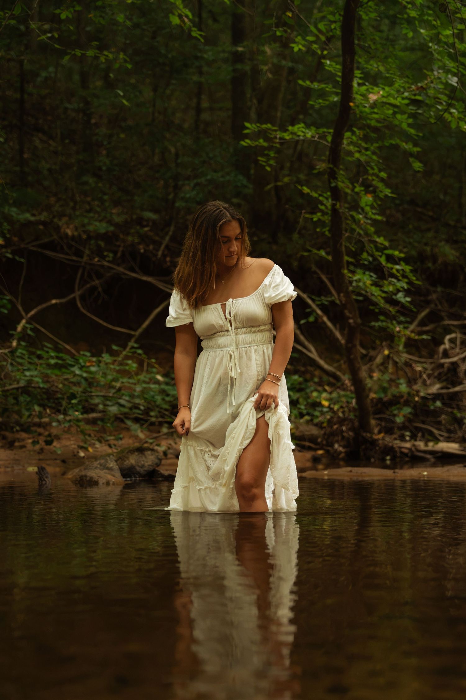 Young woman in white dress wading in forest creek, surrounded by lush green trees.
