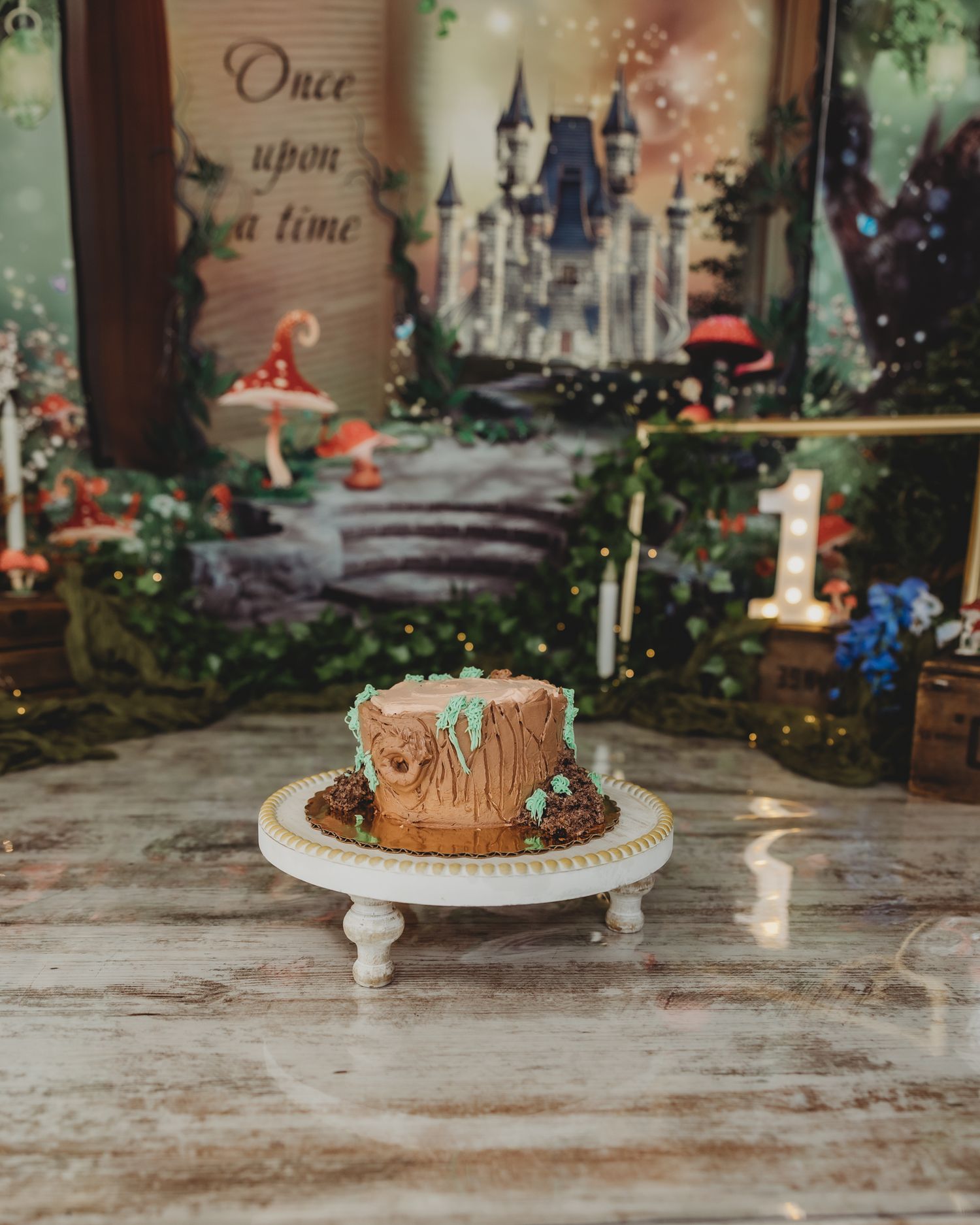 Enchanted forest themed first birthday cake on white stand with fairy tale castle backdrop and mushrooms.