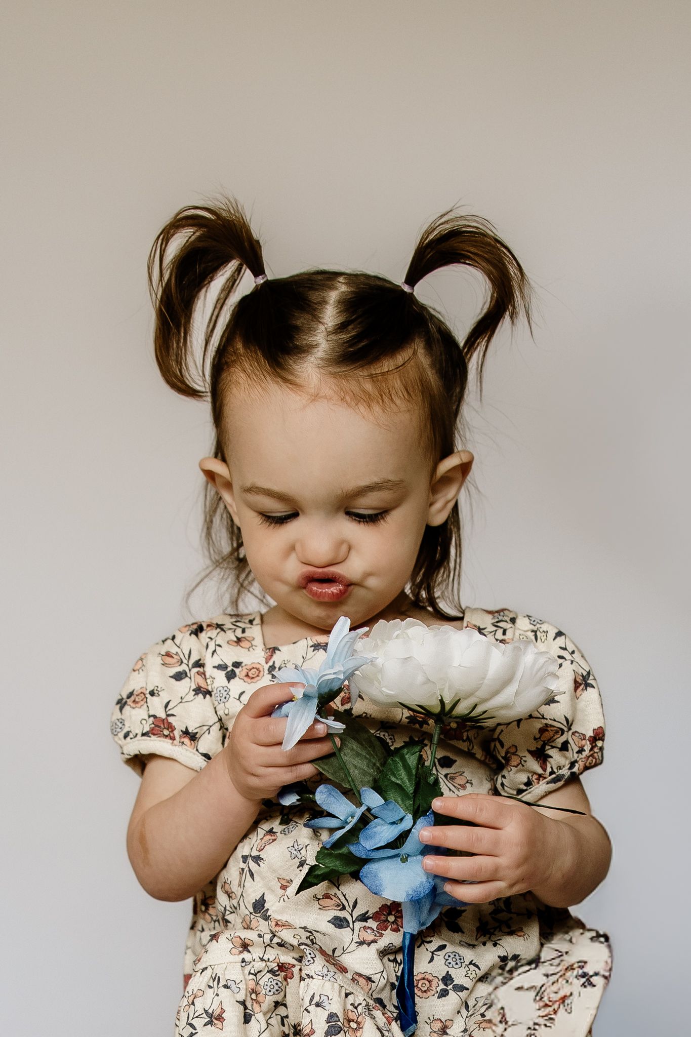 Cute toddler girl with pigtails wearing floral dress holds white and blue flower bouquet indoors.