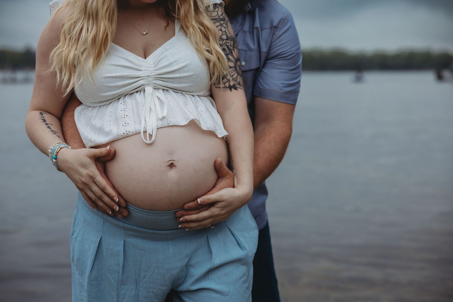A couple cradles a pregnant belly together near a lake during a maternity session.
