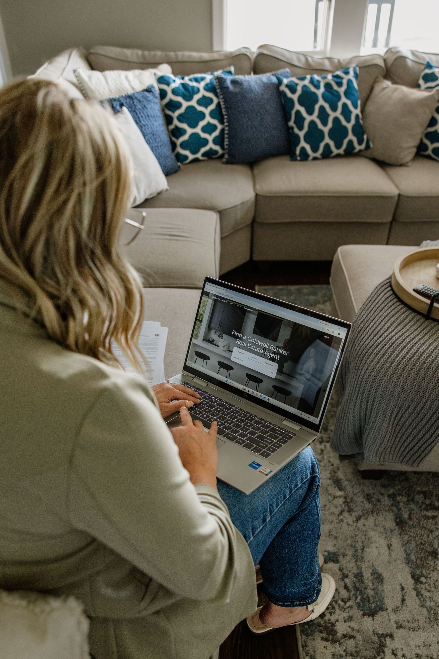 Blonde woman in olive blazer works on laptop while sitting on floor in cozy living room.