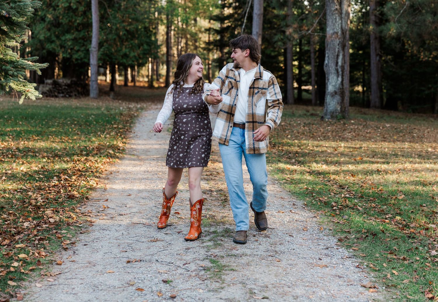 Couple walking hand-in-hand on a forest path in autumn, woman in floral dress with cowboy boots, man in plaid jacket.