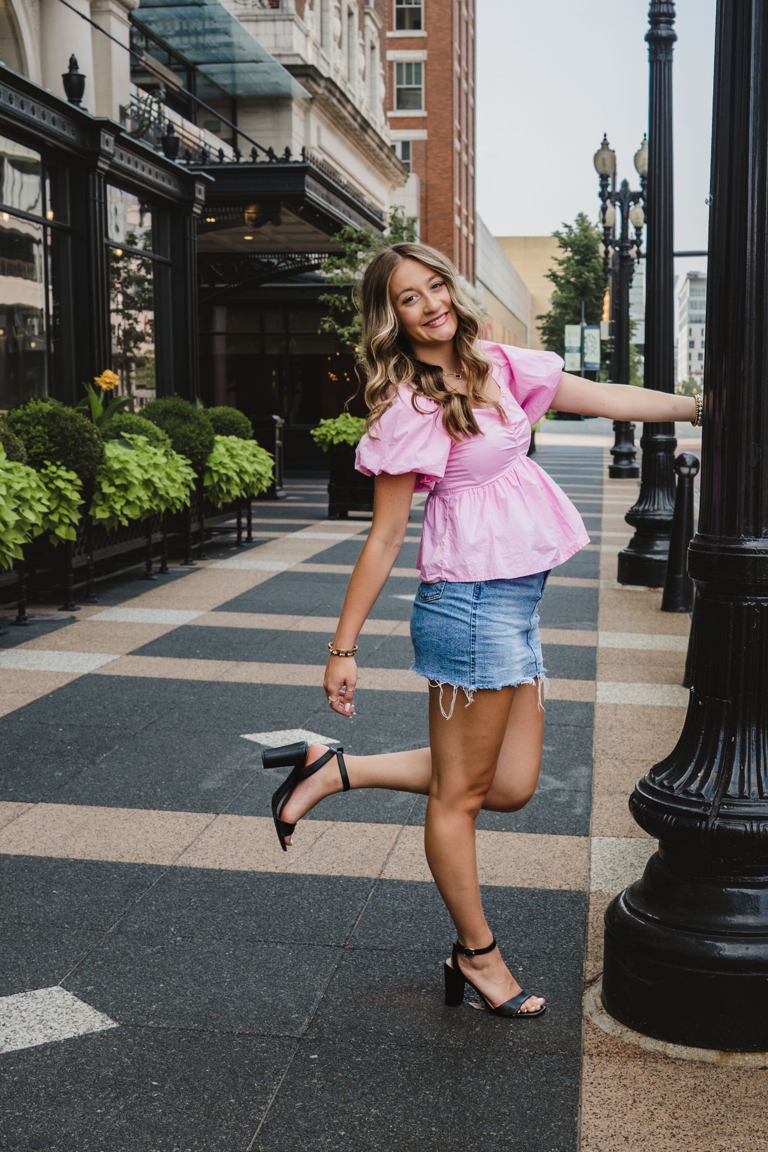 Woman in pink peplum top and denim skirt posing playfully on urban sidewalk, holding lamppost with one heel raised.