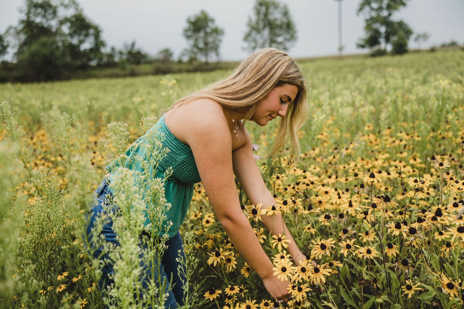 A blonde woman in a floral dress picks yellow wildflowers in a lush green meadow field.