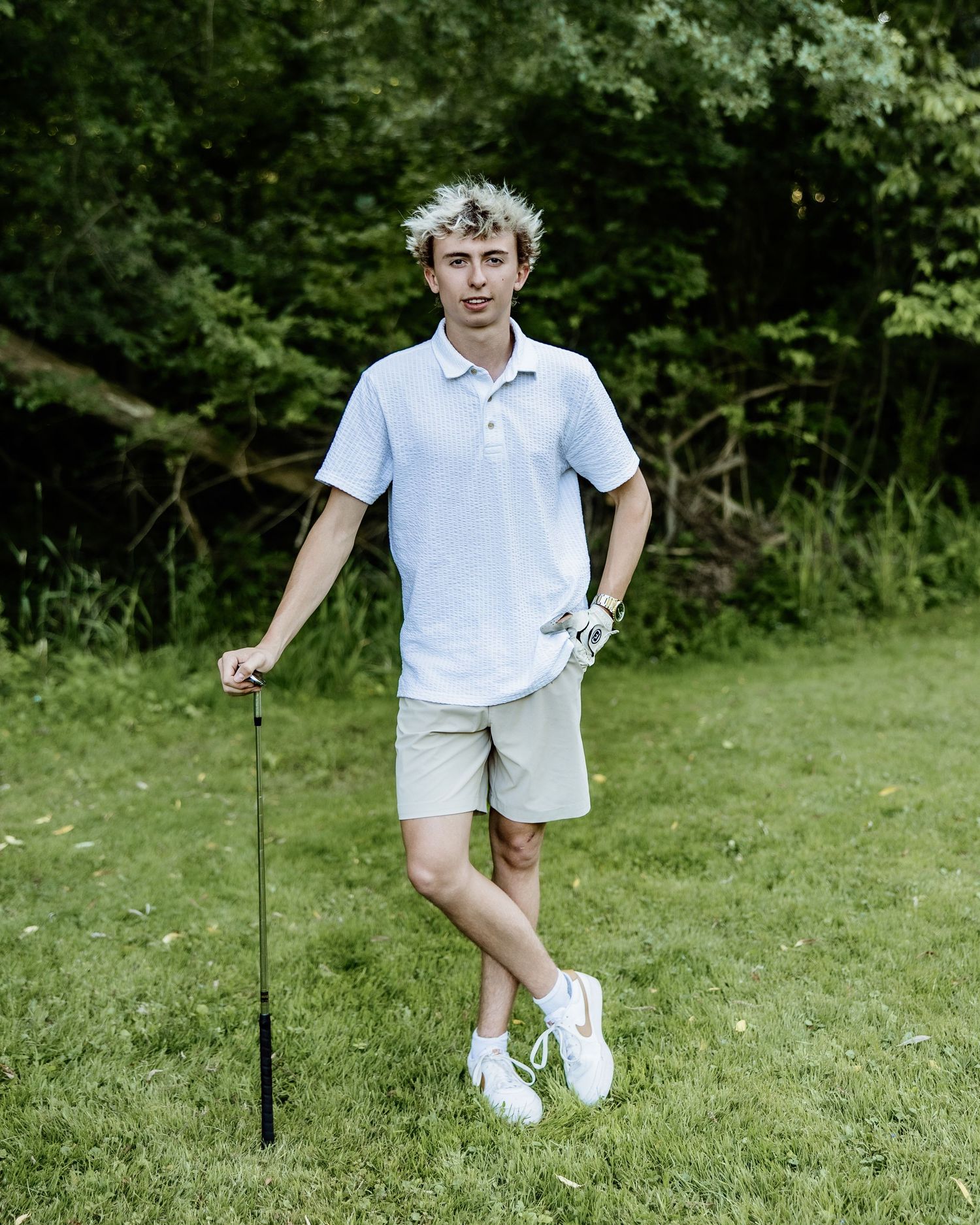 Young man in light blue polo and khaki shorts holding a golf club on a lush green golf course.