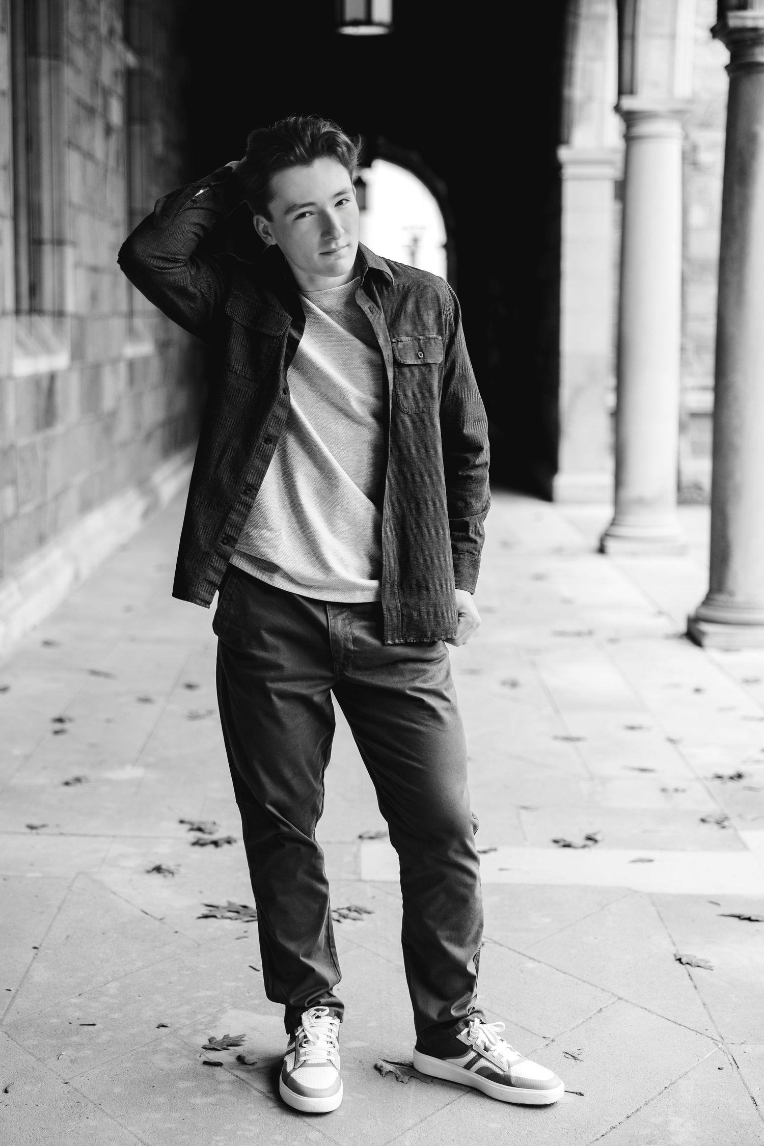 Black and white photo of a young man in jeans and jacket posing in a historic stone colonnade.