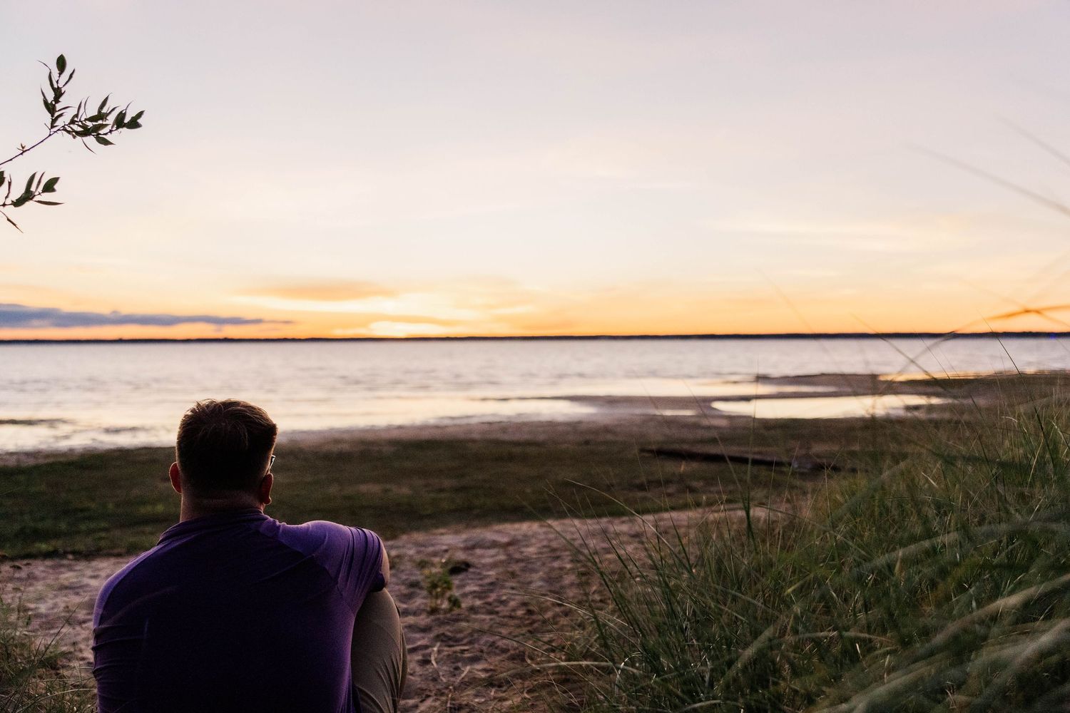 Man sitting alone on sandy shore watching colorful sunset over calm lake surrounded by tall grass.