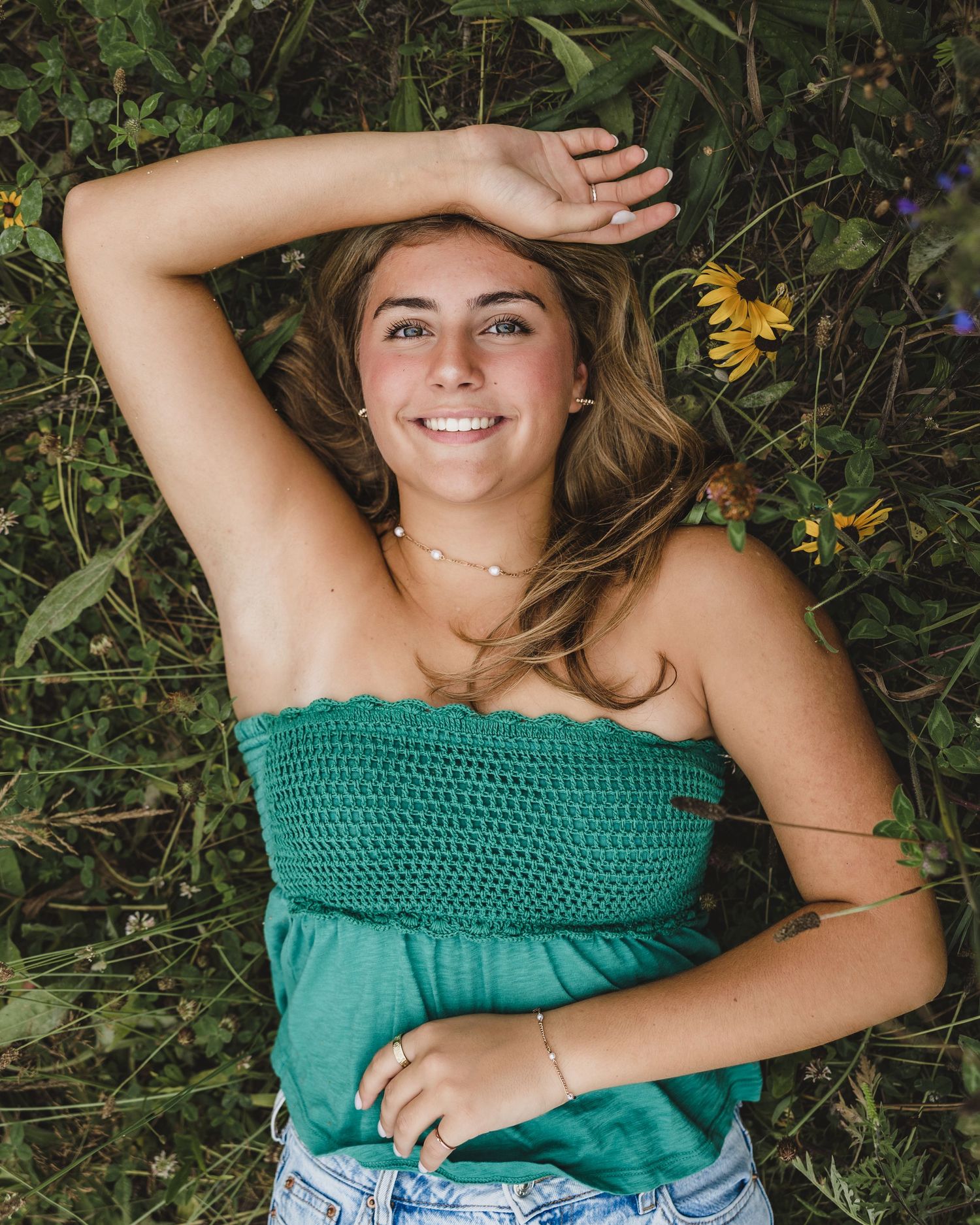 Young woman in green tube top lying in wildflower field, smiling with arm raised, wearing delicate necklace.
