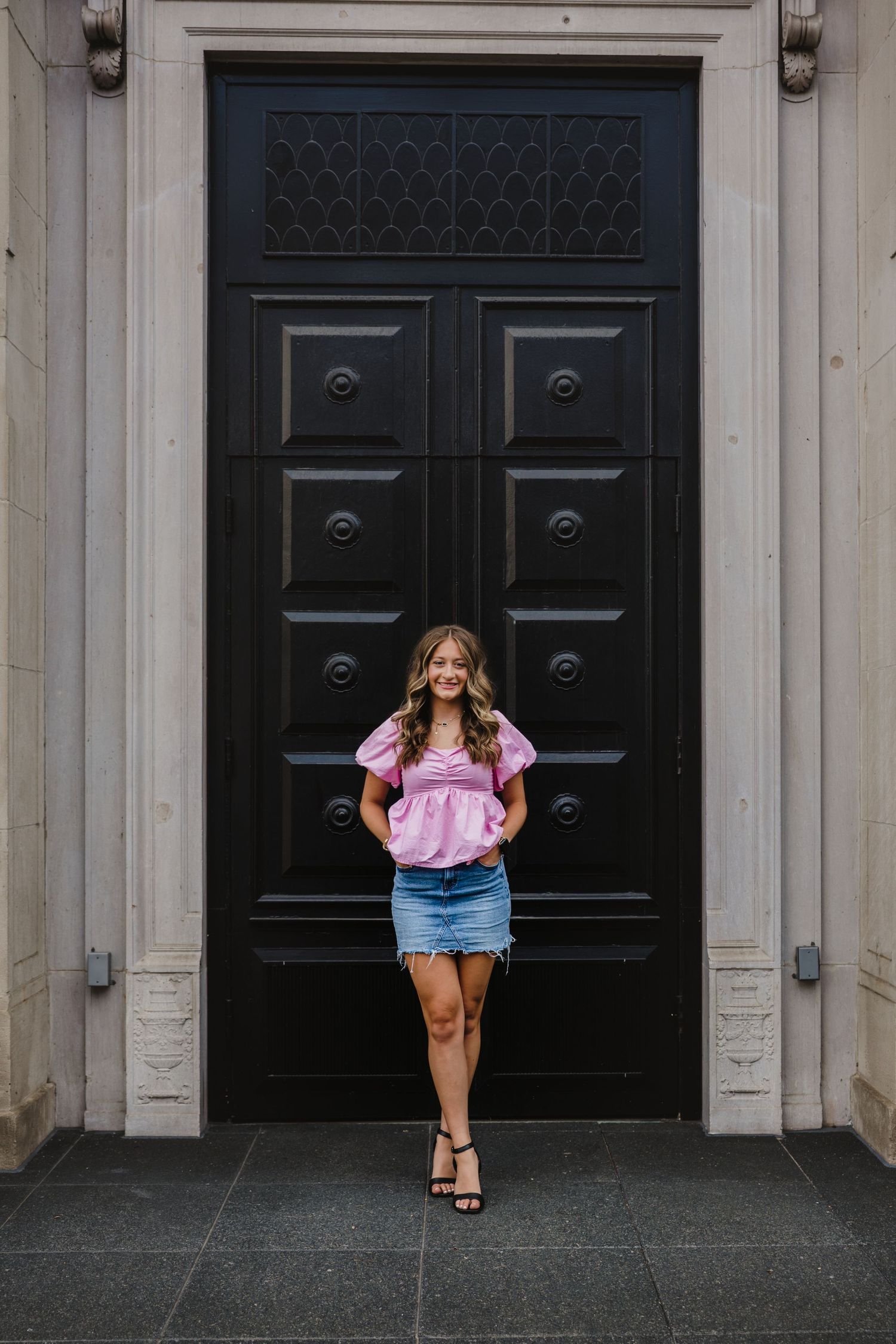 Young woman in pink top and denim skirt smiling in front of large black ornate door.