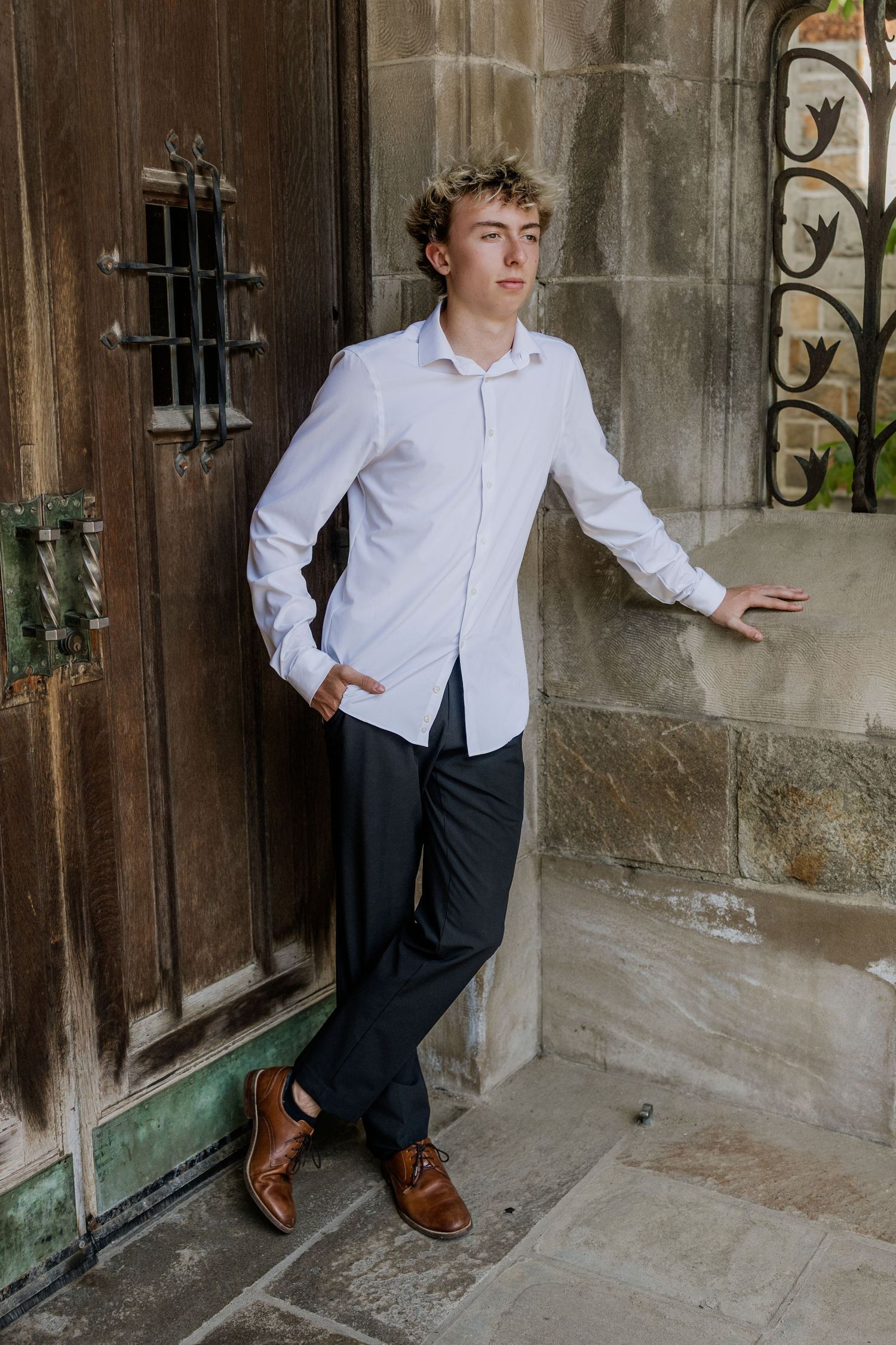 Young man in white dress shirt and dark trousers leaning against stone building with ornate wooden door.