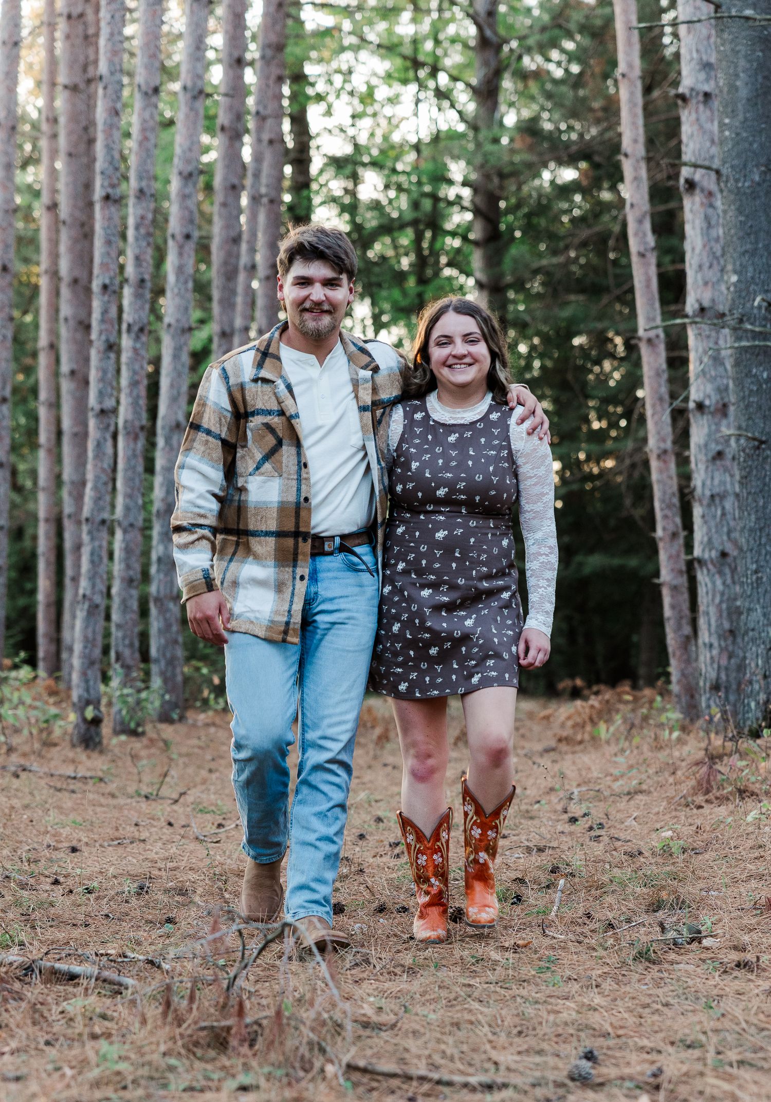 A smiling couple walks through a pine forest, man in flannel shirt and jeans, woman in floral dress and cowboy boots.