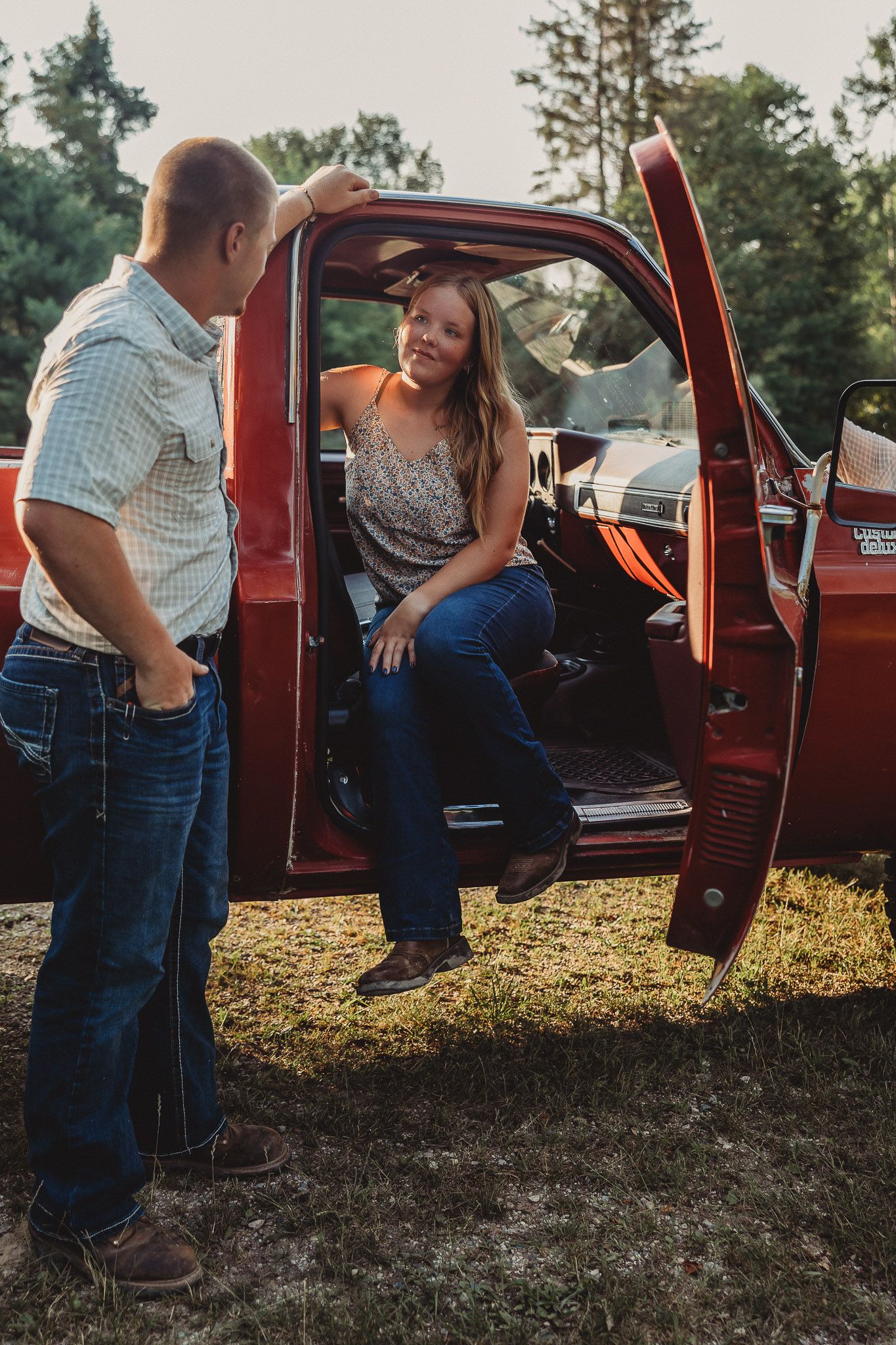 Young couple sharing a moment by an open red vintage truck door in a sunny wooded setting.