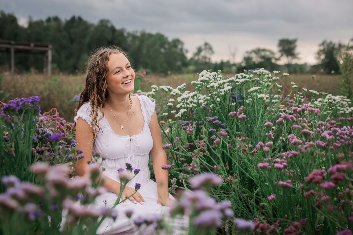 Young woman in white dress sitting among colorful wildflowers in a field, smiling joyfully on a cloudy day.