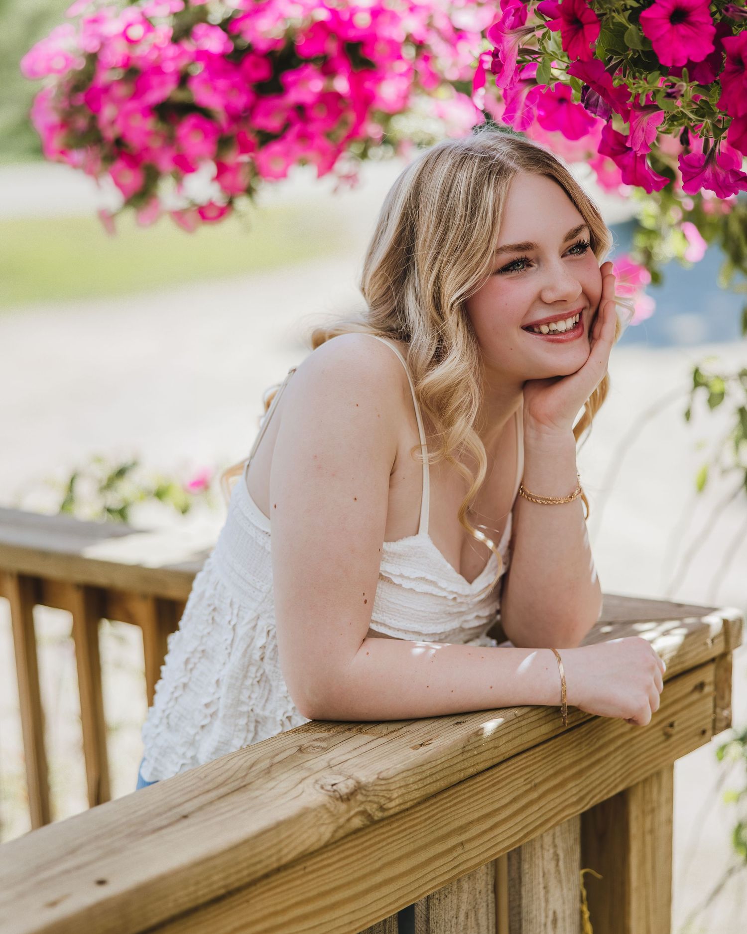Smiling blonde woman in white dress leaning on wooden railing surrounded by vibrant pink petunia flowers.