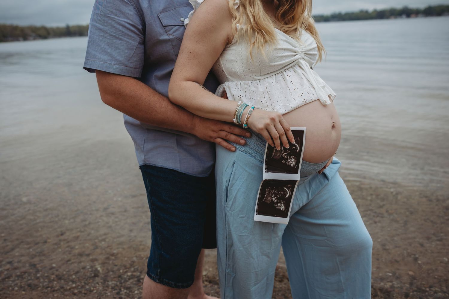 A couple holds ultrasound photos while standing by a lake during a maternity shoot.