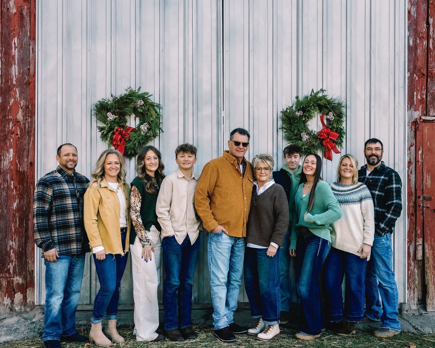 A large family of ten smiles together outside a rustic red barn with holiday wreaths.