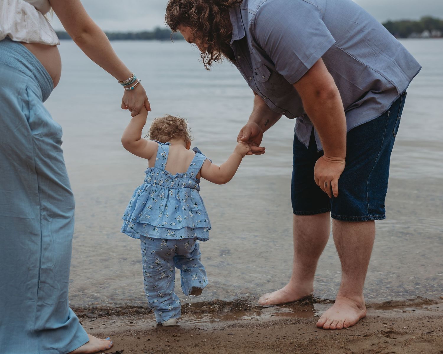 Parents holding hands with their baby girl in a blue dress at the waters edge.