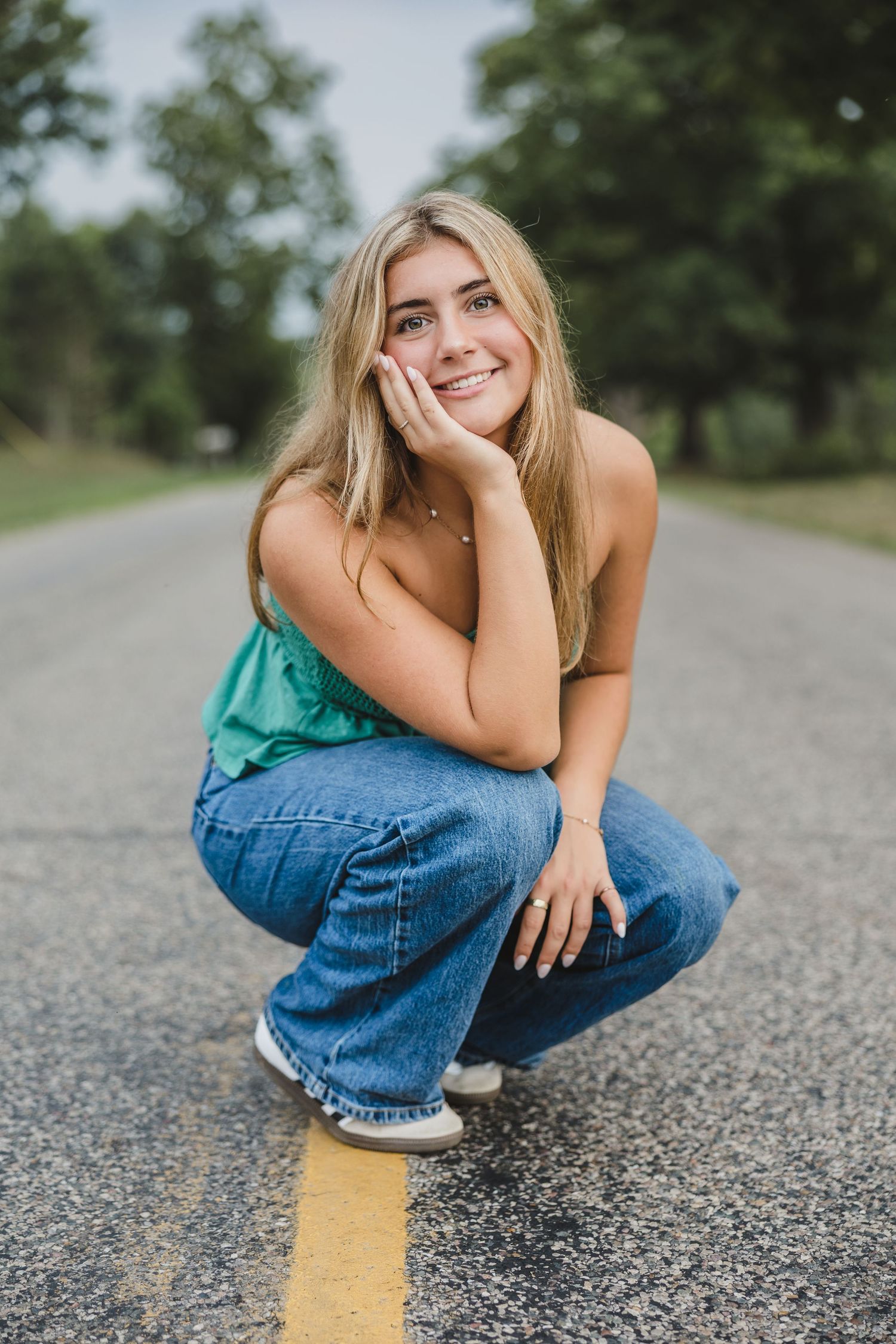 Young blonde woman in green top and jeans crouching on a road, smiling with hand on cheek.