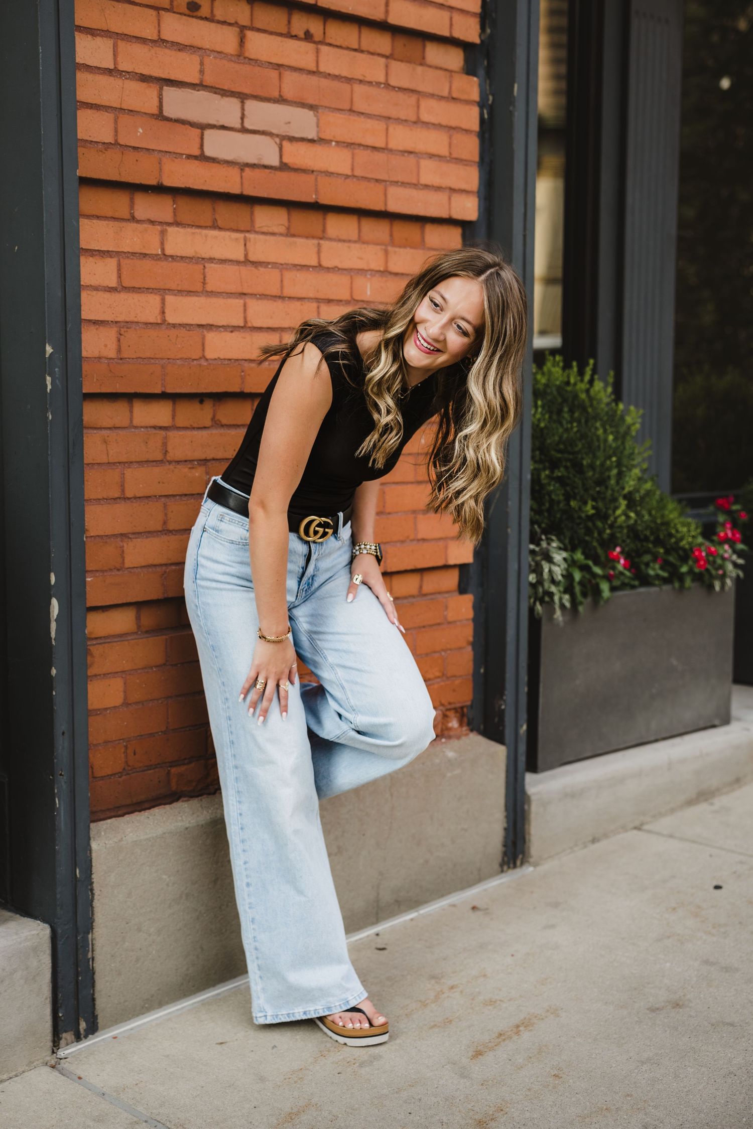 Woman in black top, wide-leg jeans, and Gucci belt posing against a brick wall outdoors.