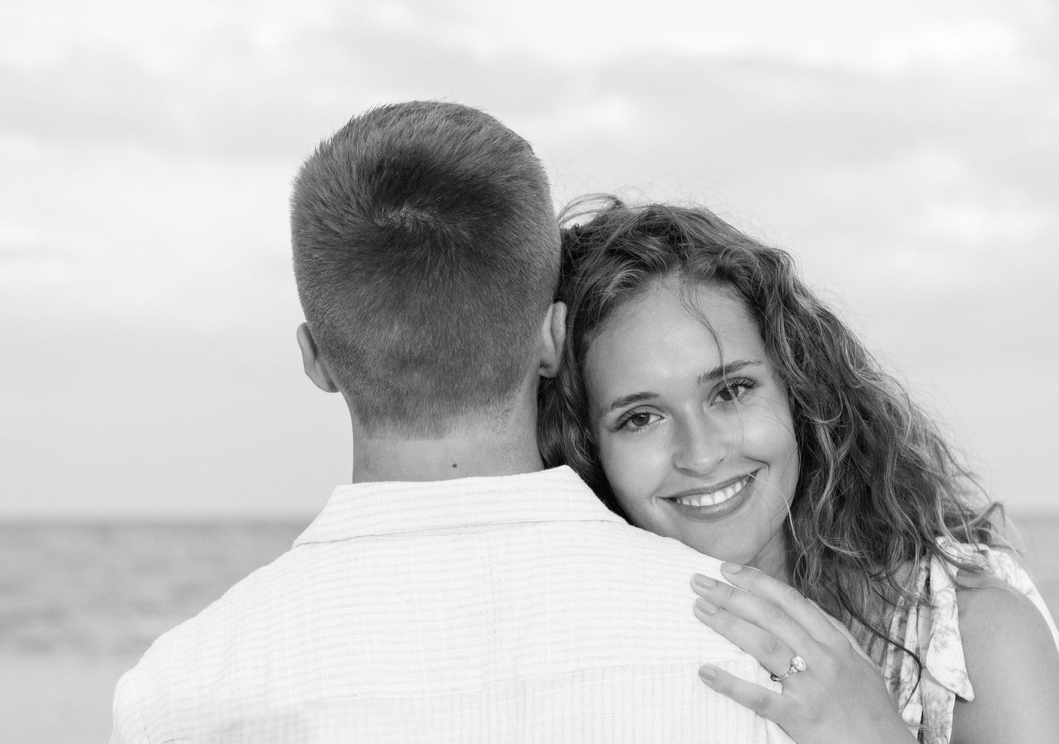 Black and white photo of a smiling woman embracing a man at the beach, showing an engagement ring.