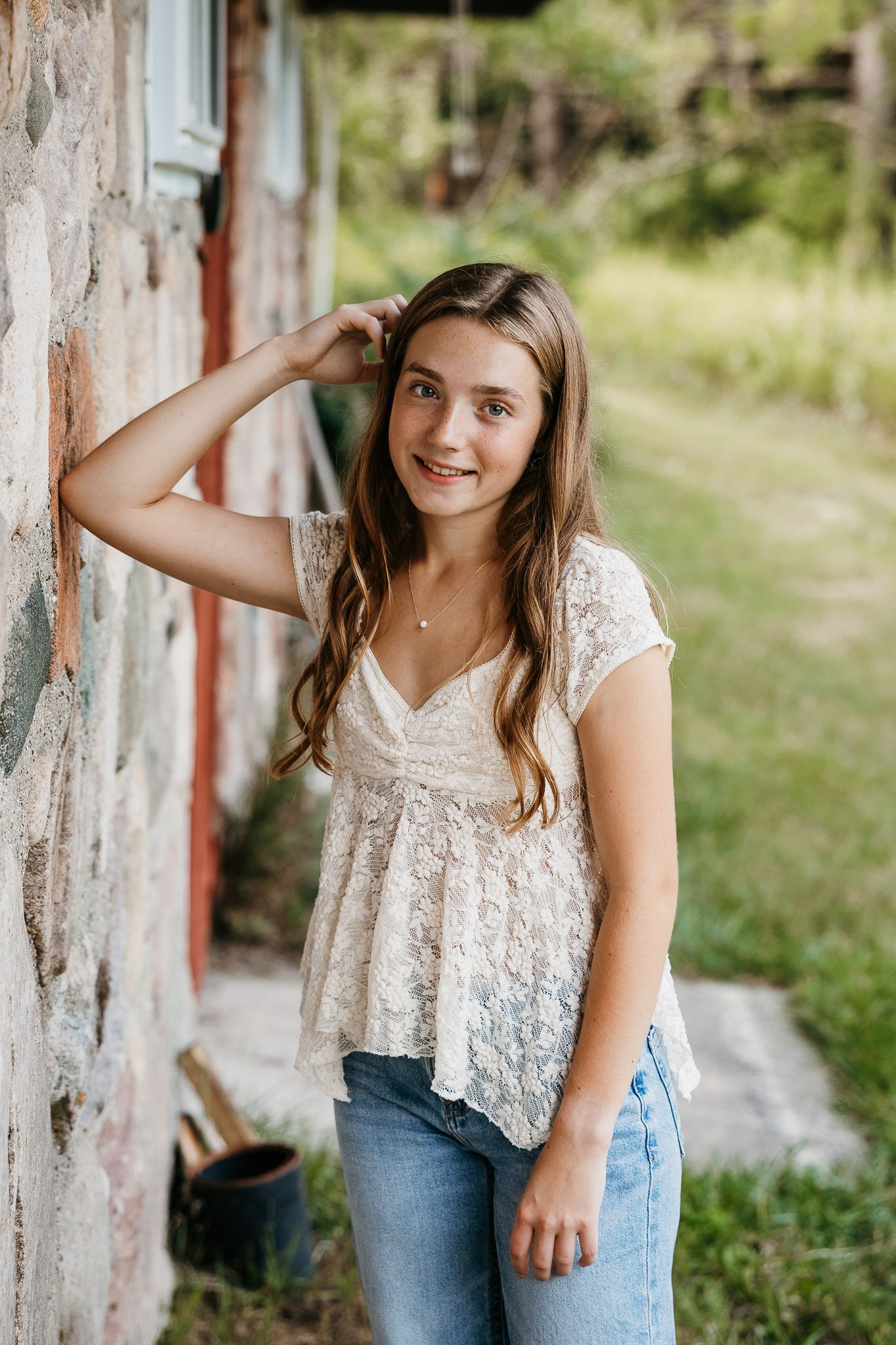 Smiling teenage girl in white lace top and jeans leaning against a rustic stone wall outdoors.