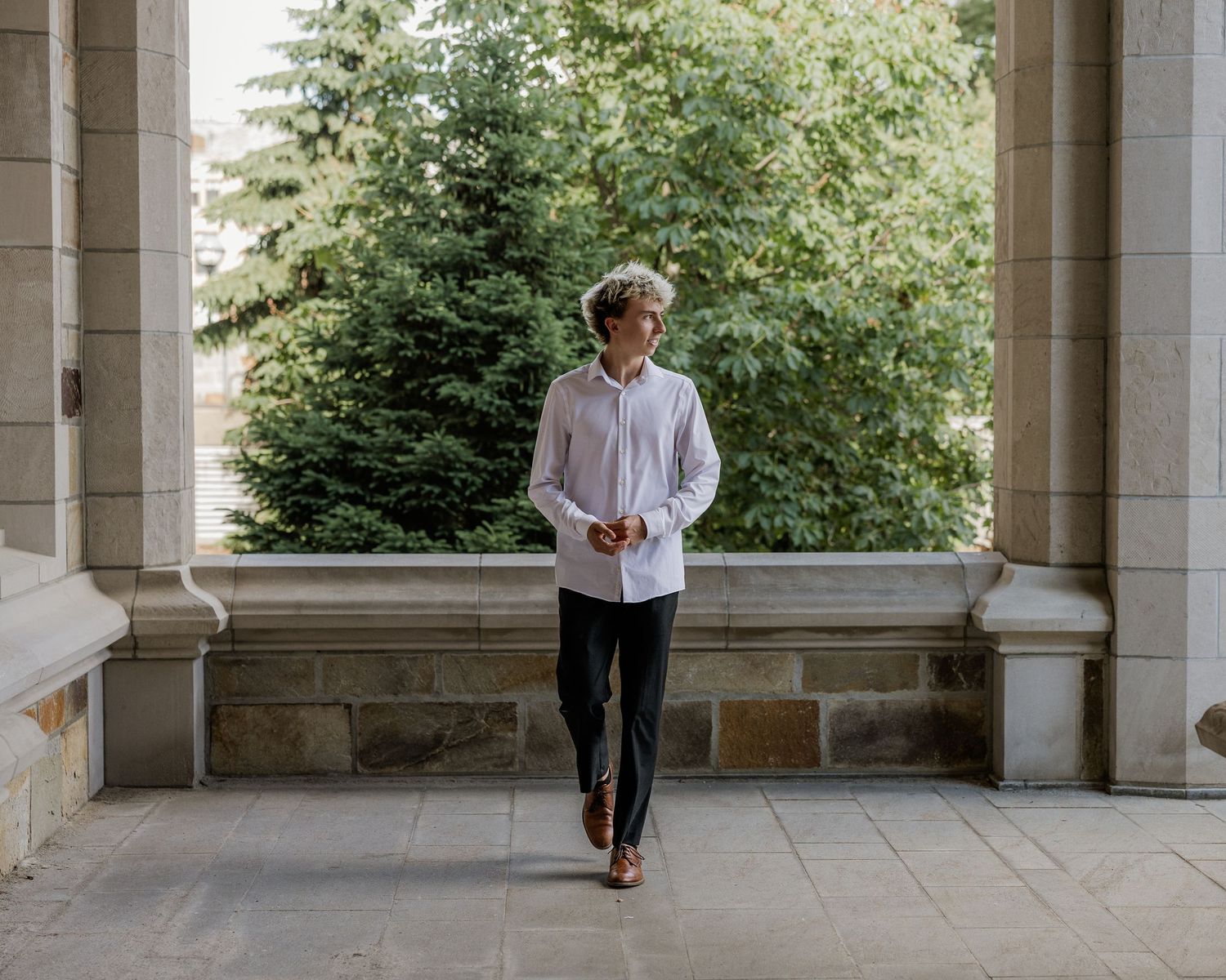 Young man in white shirt and black pants walking through stone colonnade with lush green trees in background.