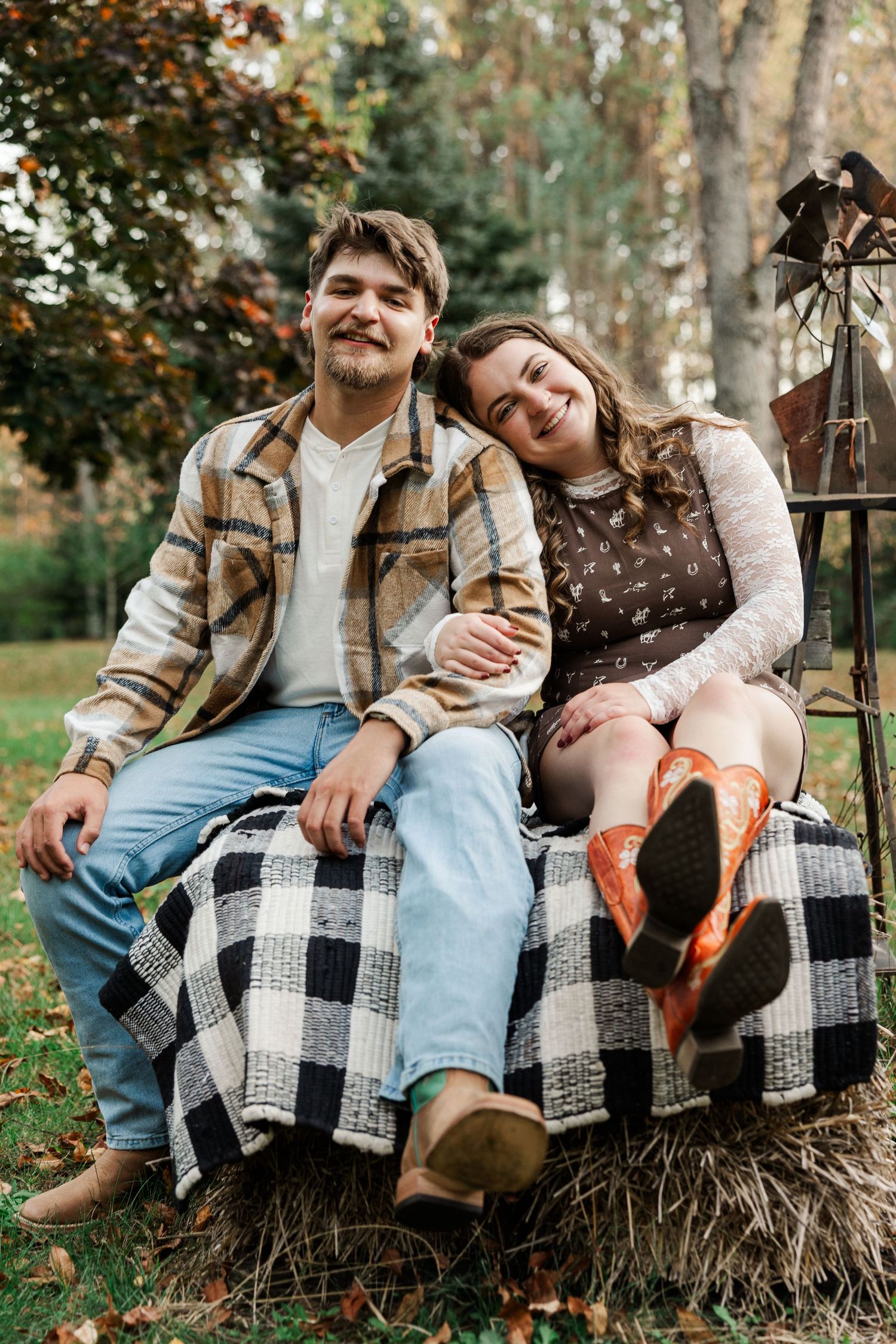 Couple sitting on hay bale with plaid blanket, smiling outdoors in autumn setting, woman wearing orange cowboy boots.