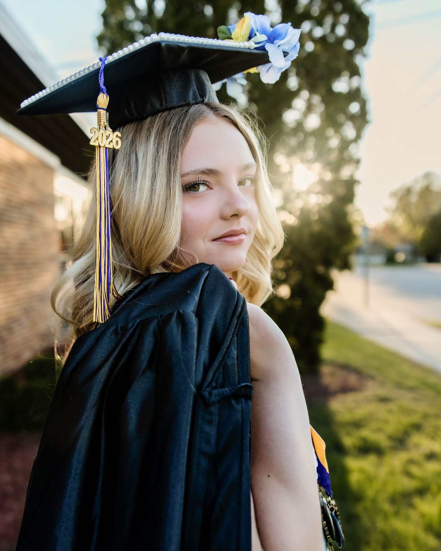 Young female graduate in black cap and gown with 2026 tassel posing outdoors in golden sunset light.