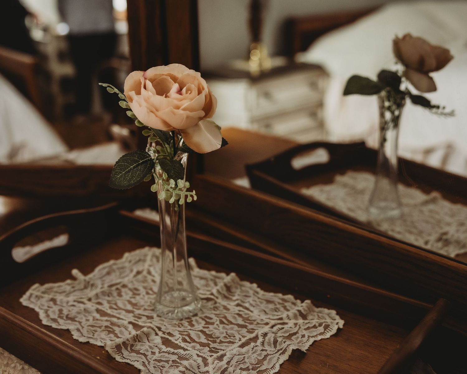 Single peach rose in glass bud vase on lace doily atop wooden serving tray.