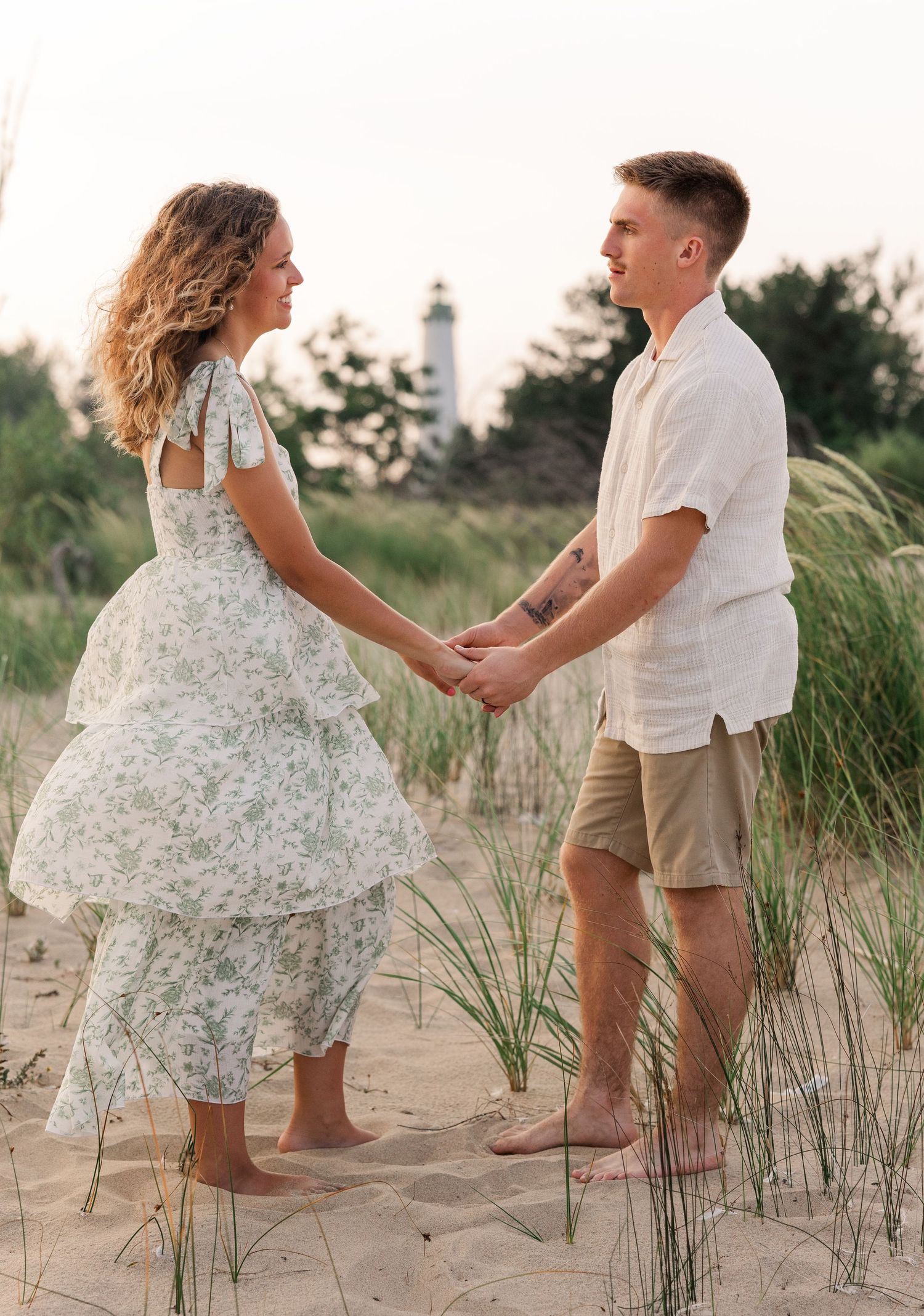 Couple holding hands barefoot on sandy beach dunes with lighthouse and trees in background at sunset.