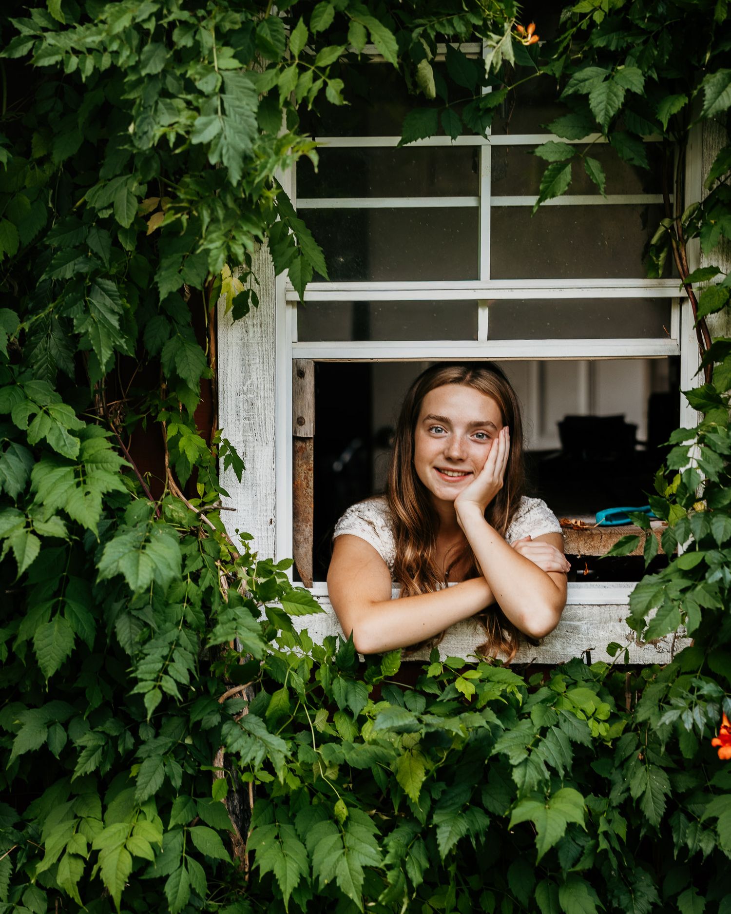 Young woman with long hair smiling out of a vine-covered white window surrounded by lush green leaves.