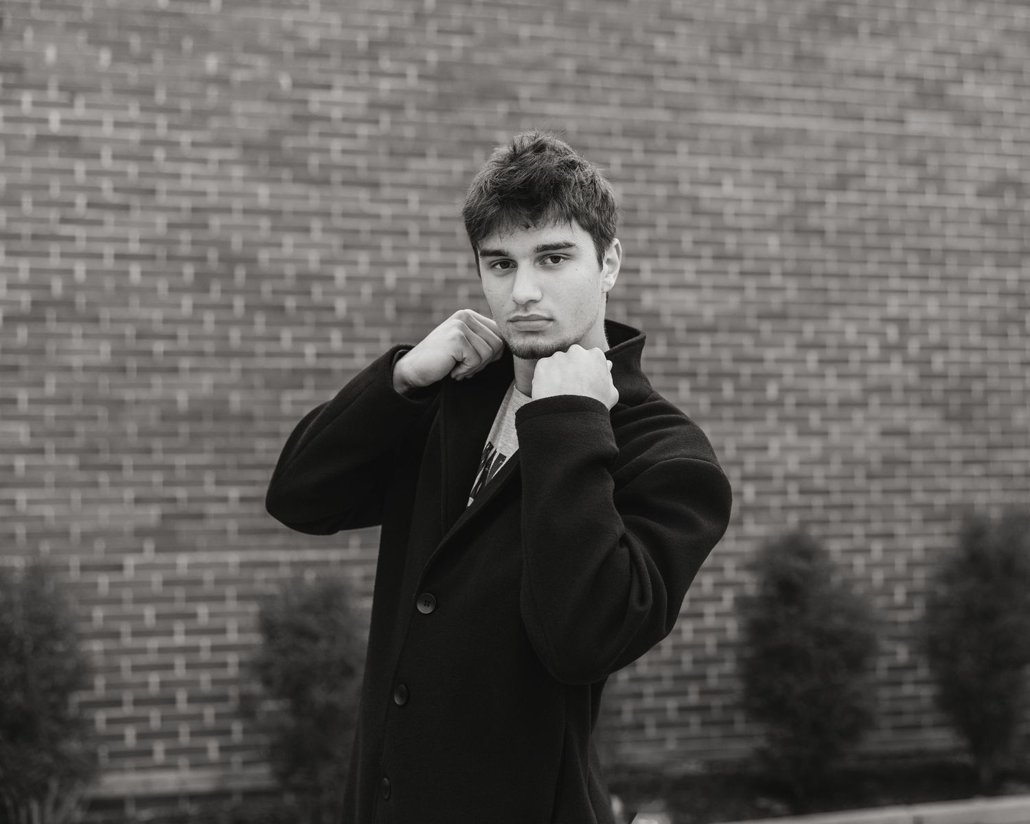 Young man in black coat poses with hands raised near chin against brick wall in black and white photo.