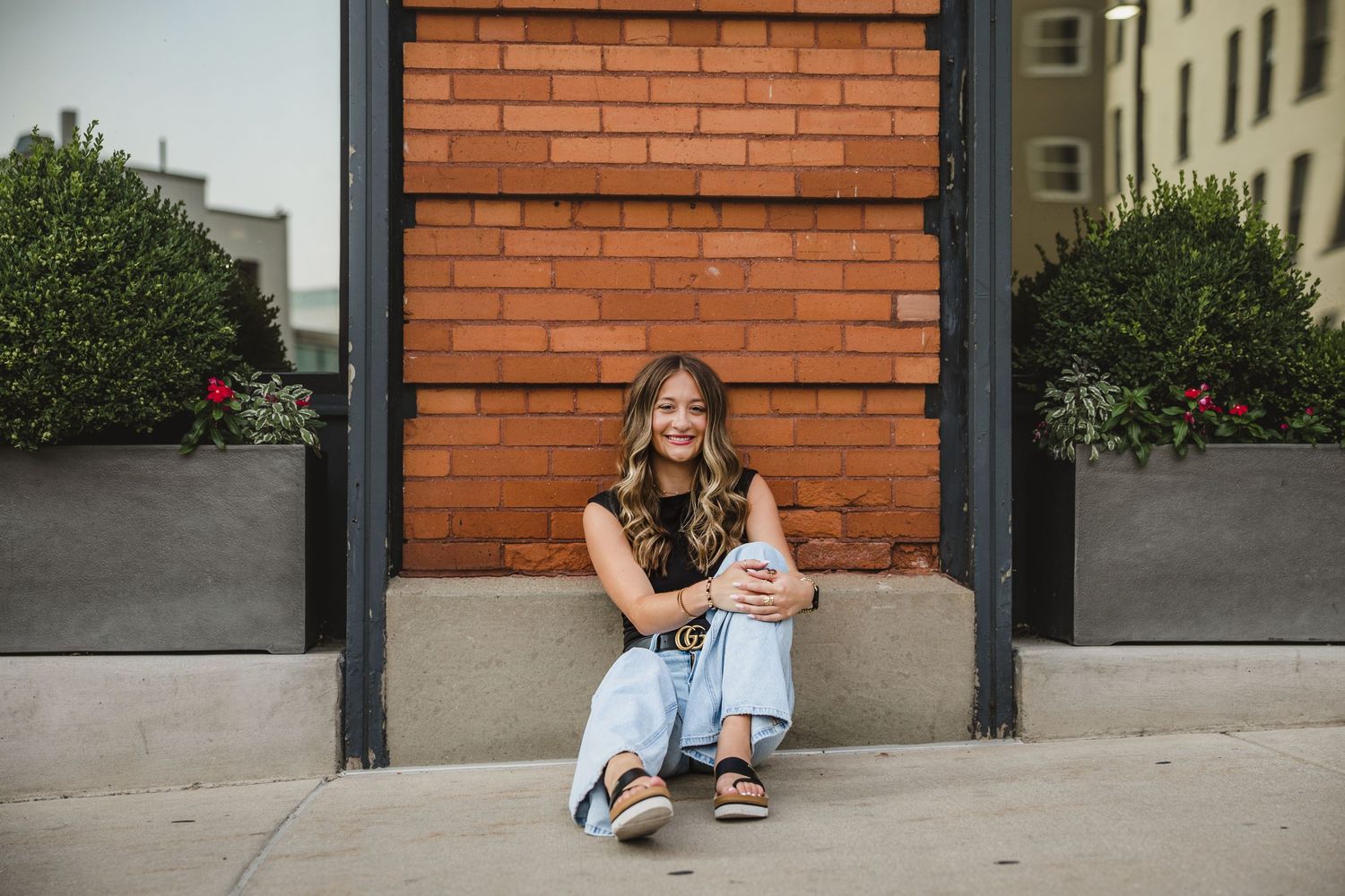 Young woman smiling, sitting on steps against a red brick wall, wearing jeans and a dark top, flanked by green planters.