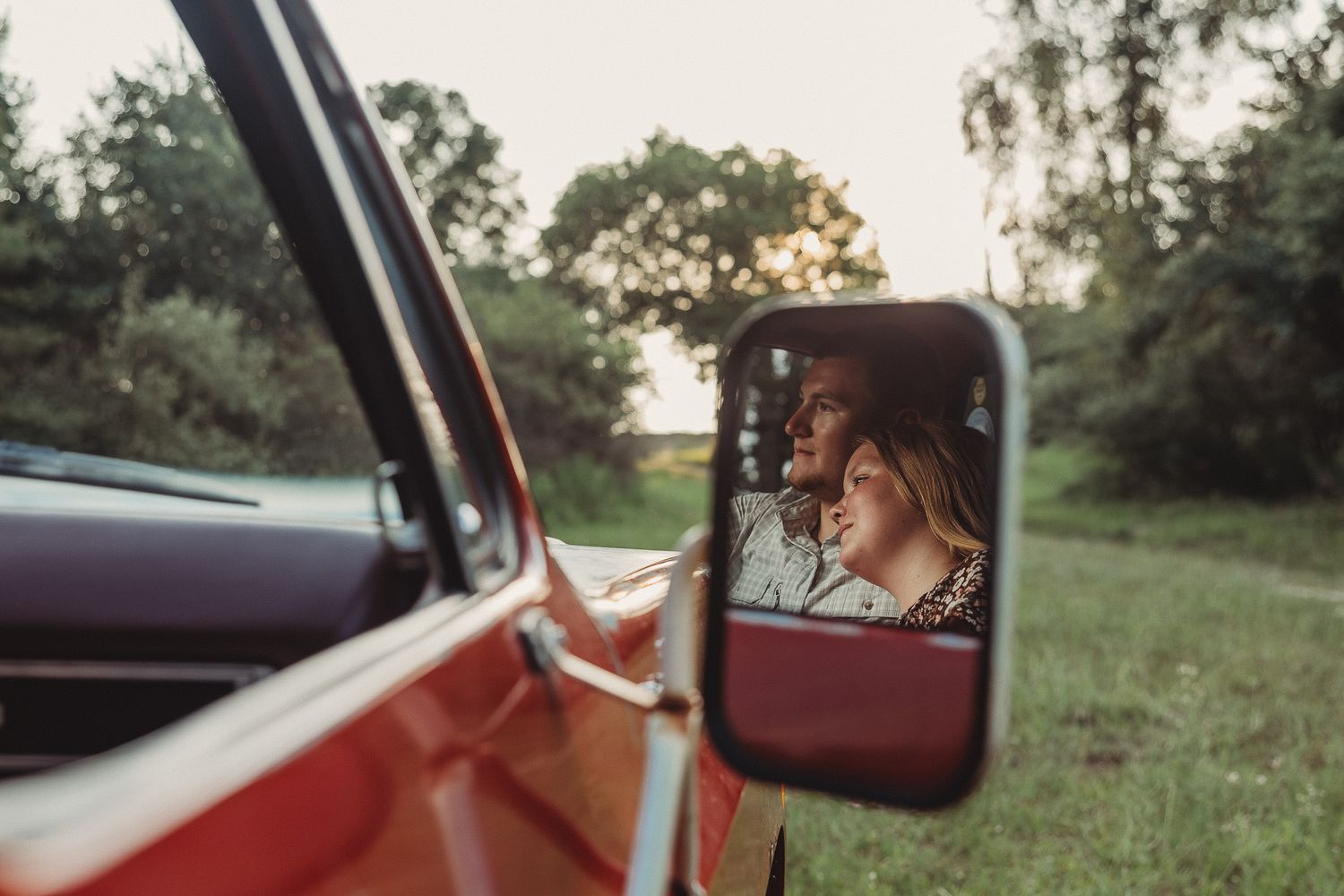 Romantic couple reflected in a vintage red truck side mirror at golden hour in a green field.
