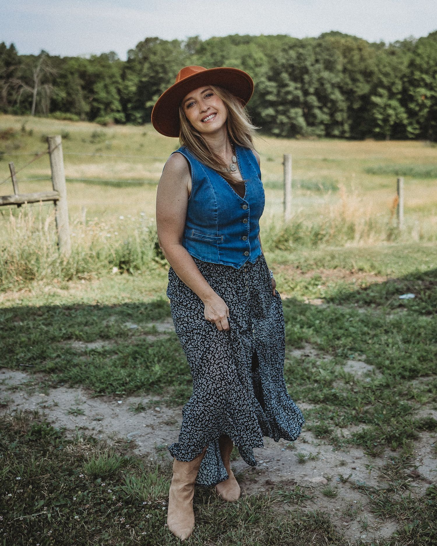 Laughing woman in rust hat, denim vest, and floral maxi skirt twirling on a sunny farm field.