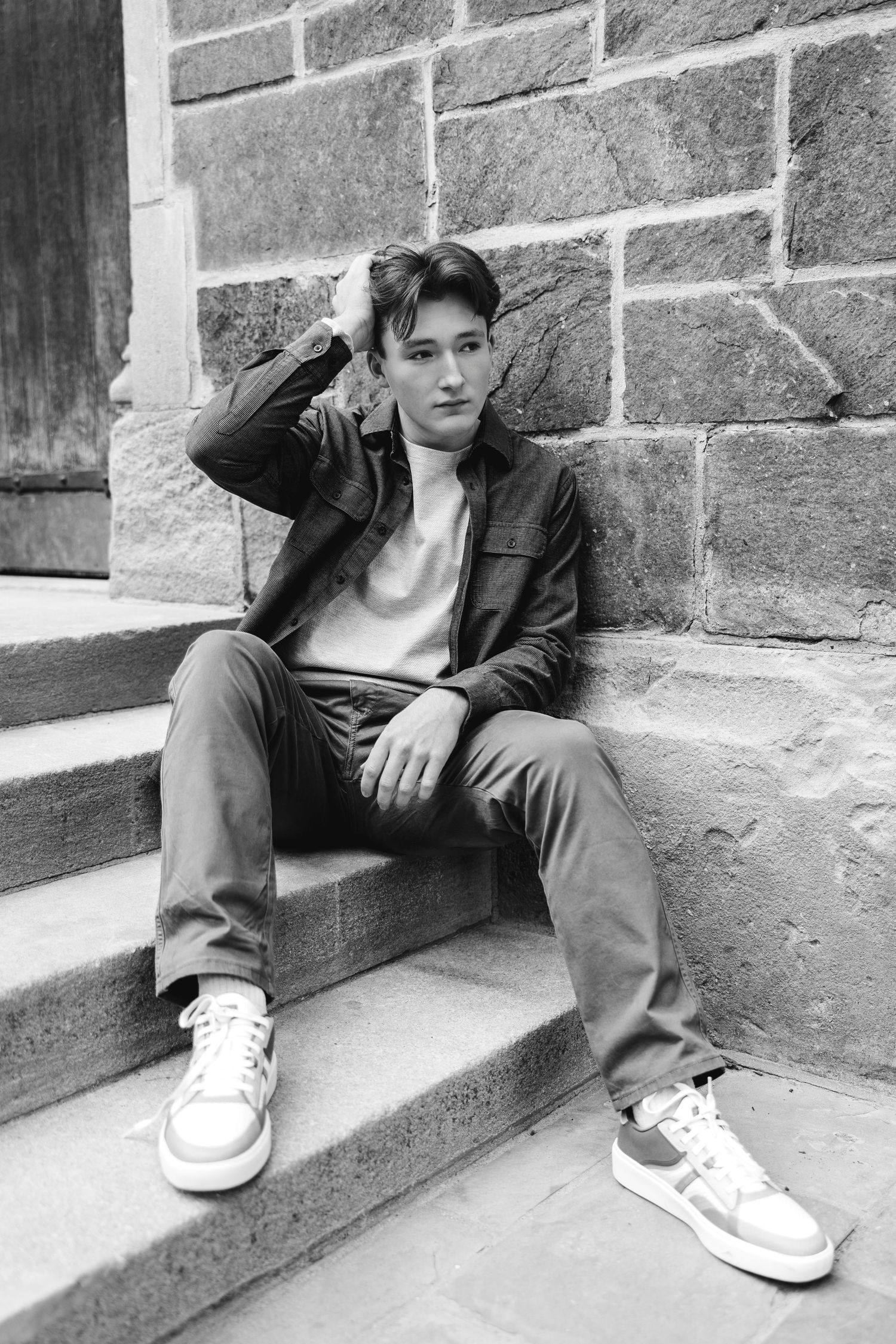 Black and white portrait of a young man sitting on stone steps leaning against a brick wall.