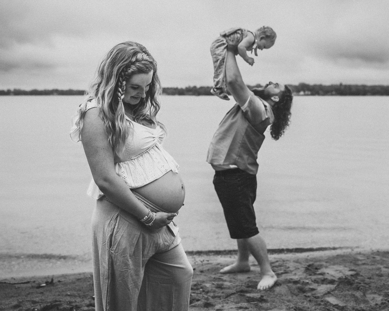 Black and white maternity photo of pregnant woman on beach while partner lifts toddler.