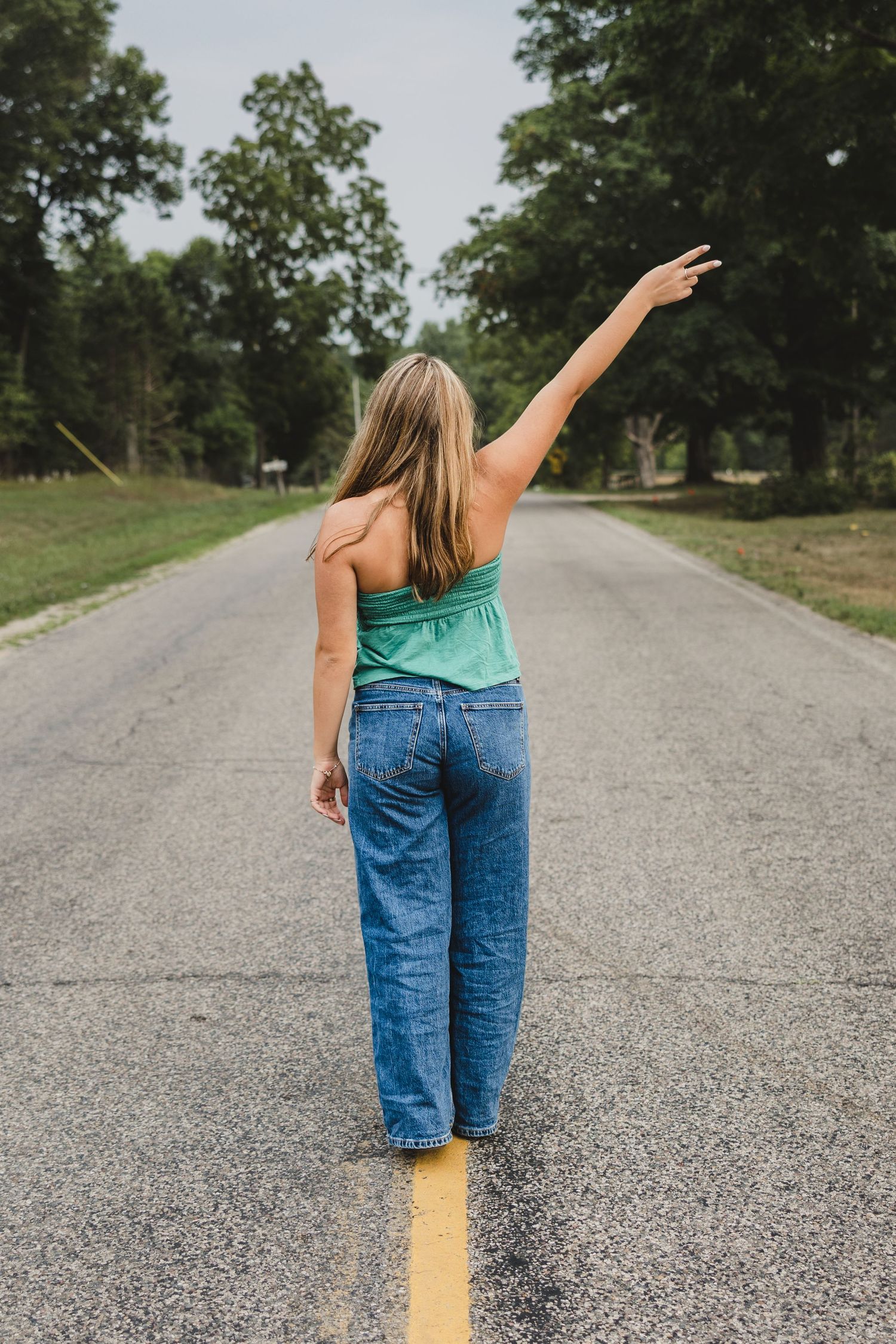 Woman in green top and jeans standing on road center line, arm raised, surrounded by tree-lined street.
