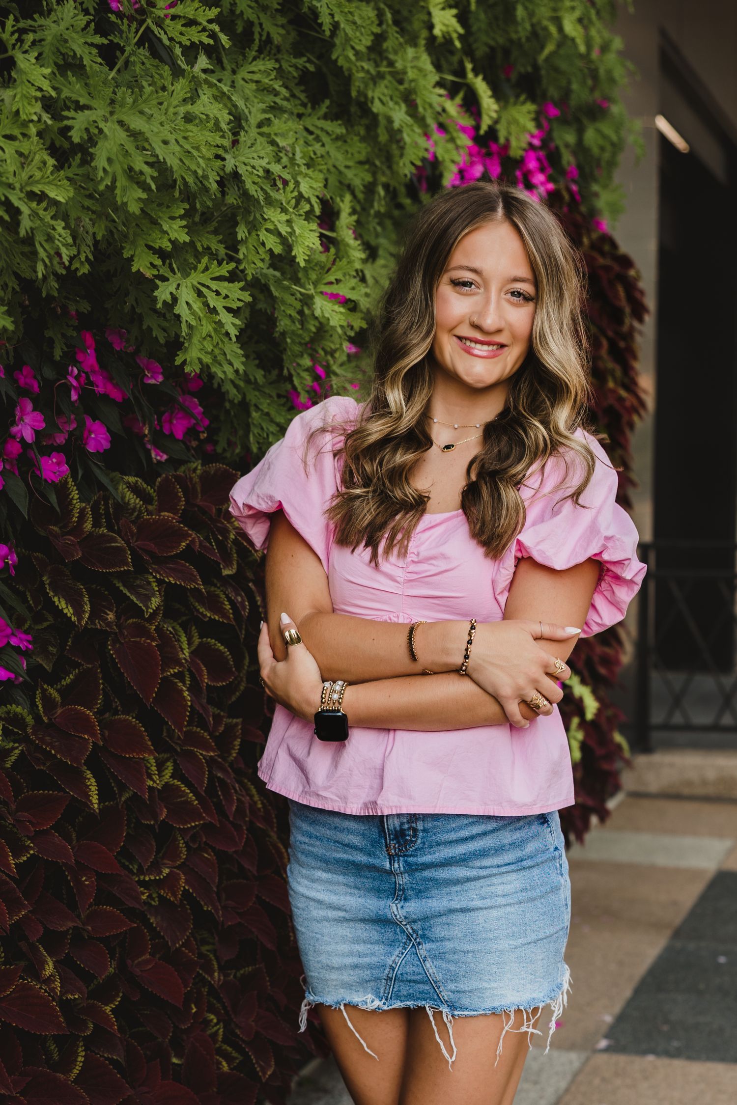 Smiling young woman in pink ruffled top and denim skirt posing outdoors near colorful flowers and foliage.