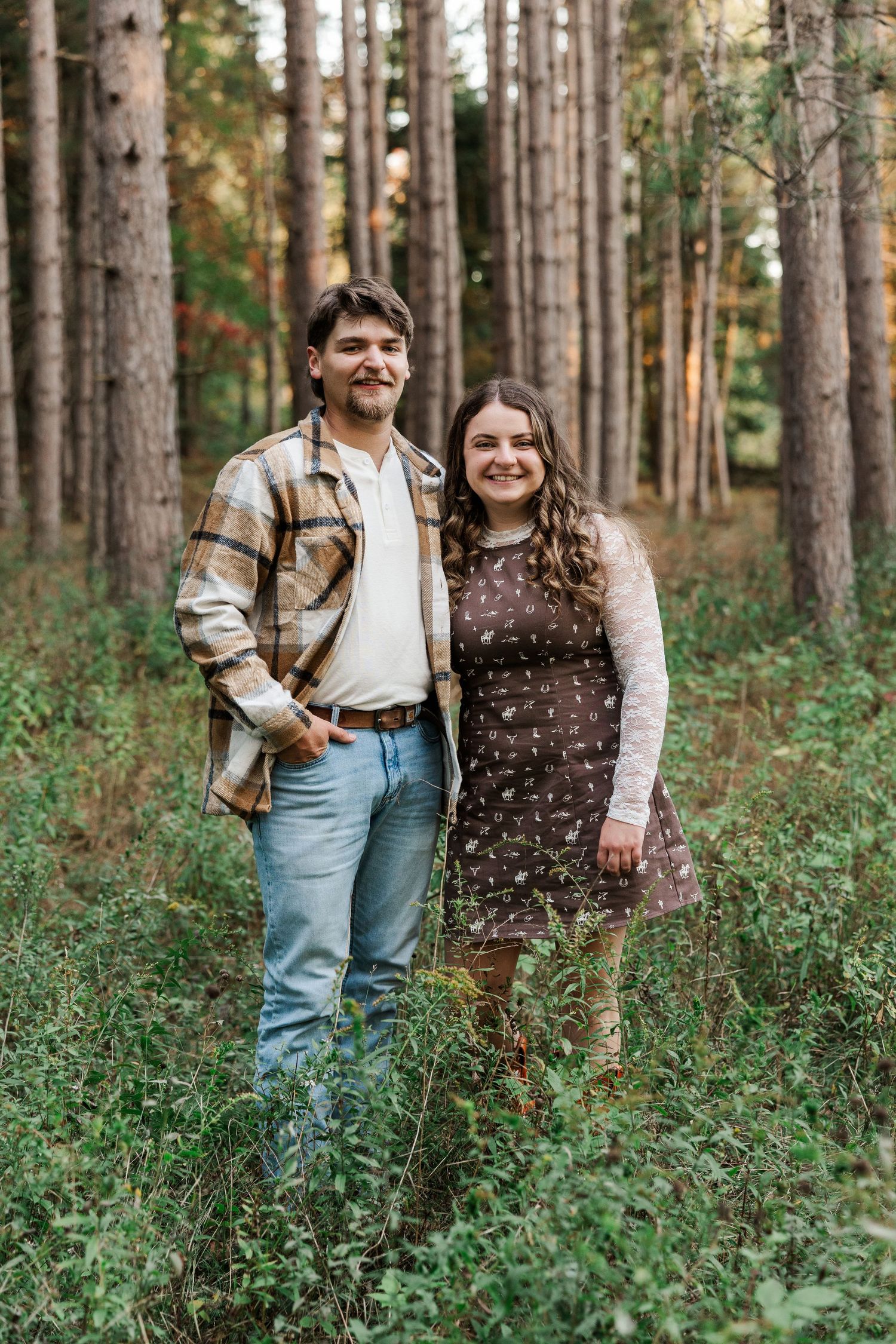 A happy couple stands together in a lush pine forest, the man in plaid and the woman in a brown dress.