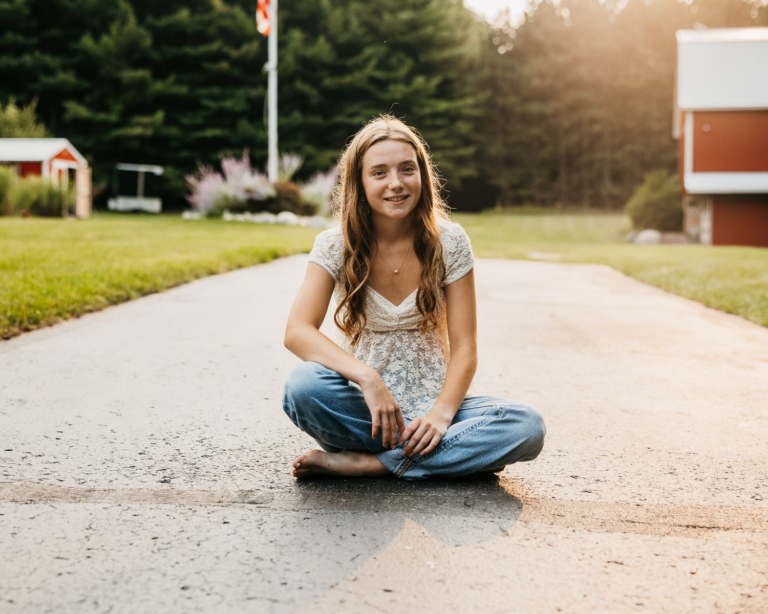 Young woman with long blonde hair sitting cross-legged on a driveway, smiling, wearing jeans and a floral top at golden hour.