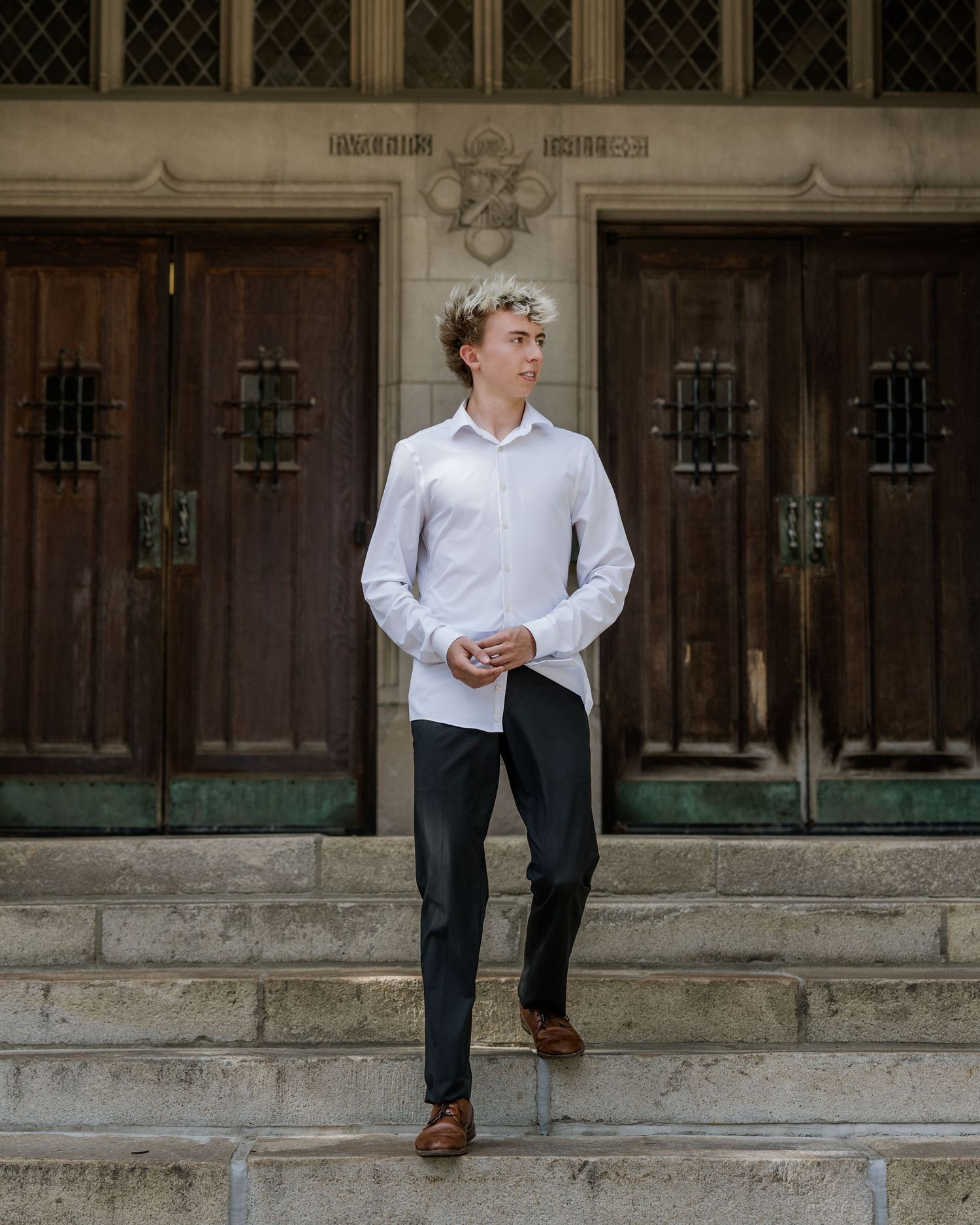 Young man in white shirt and dark trousers stands on stone steps before ornate wooden doors.