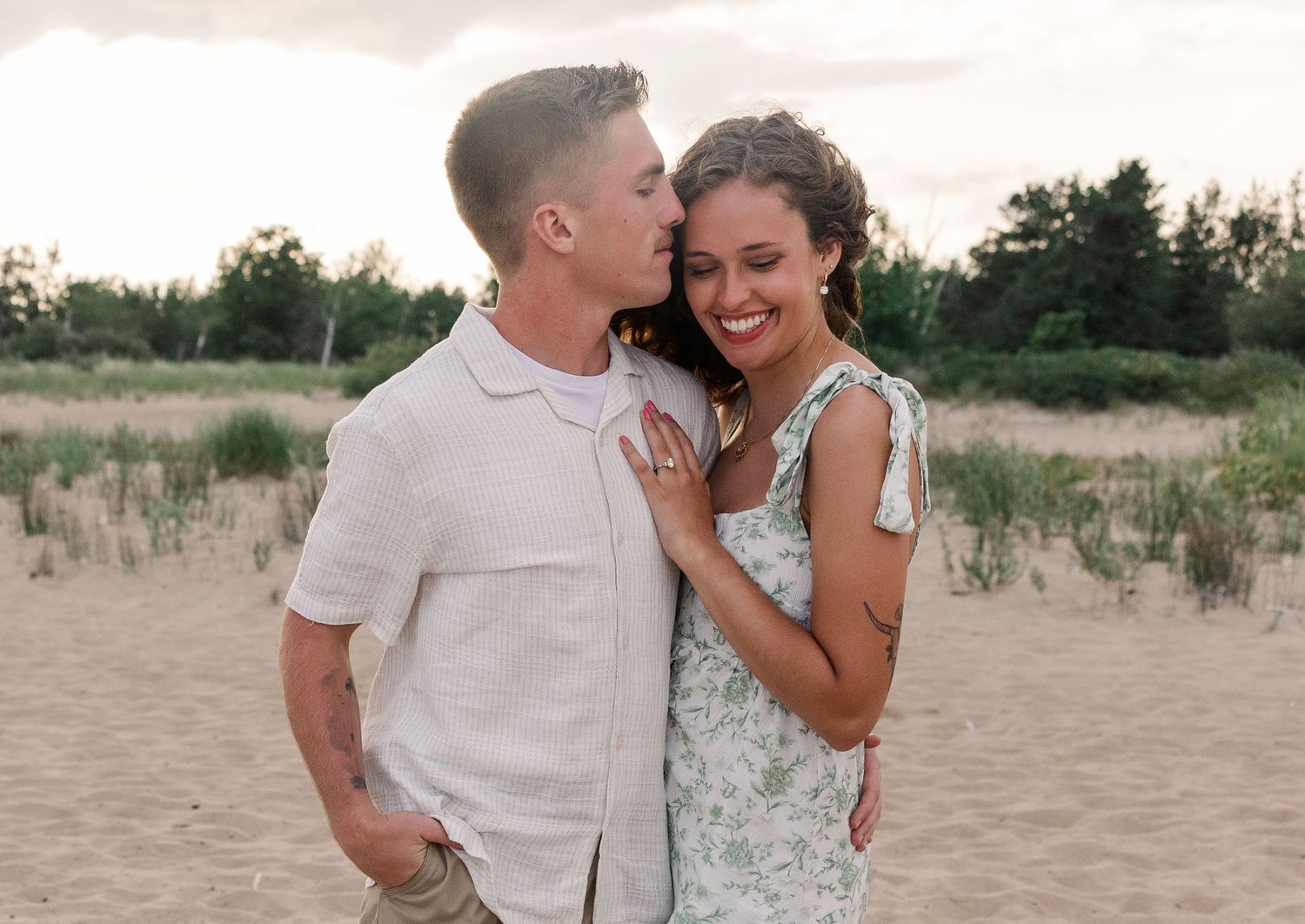 A happy couple shares a romantic moment on a sandy beach, smiling and embracing near lush green trees.