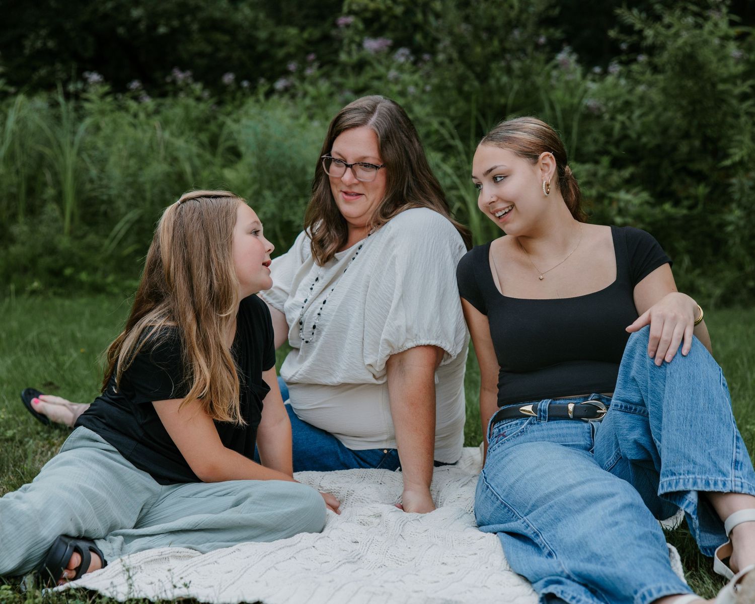 Three women sitting together on a blanket outdoors, smiling and laughing in a lush green garden setting.