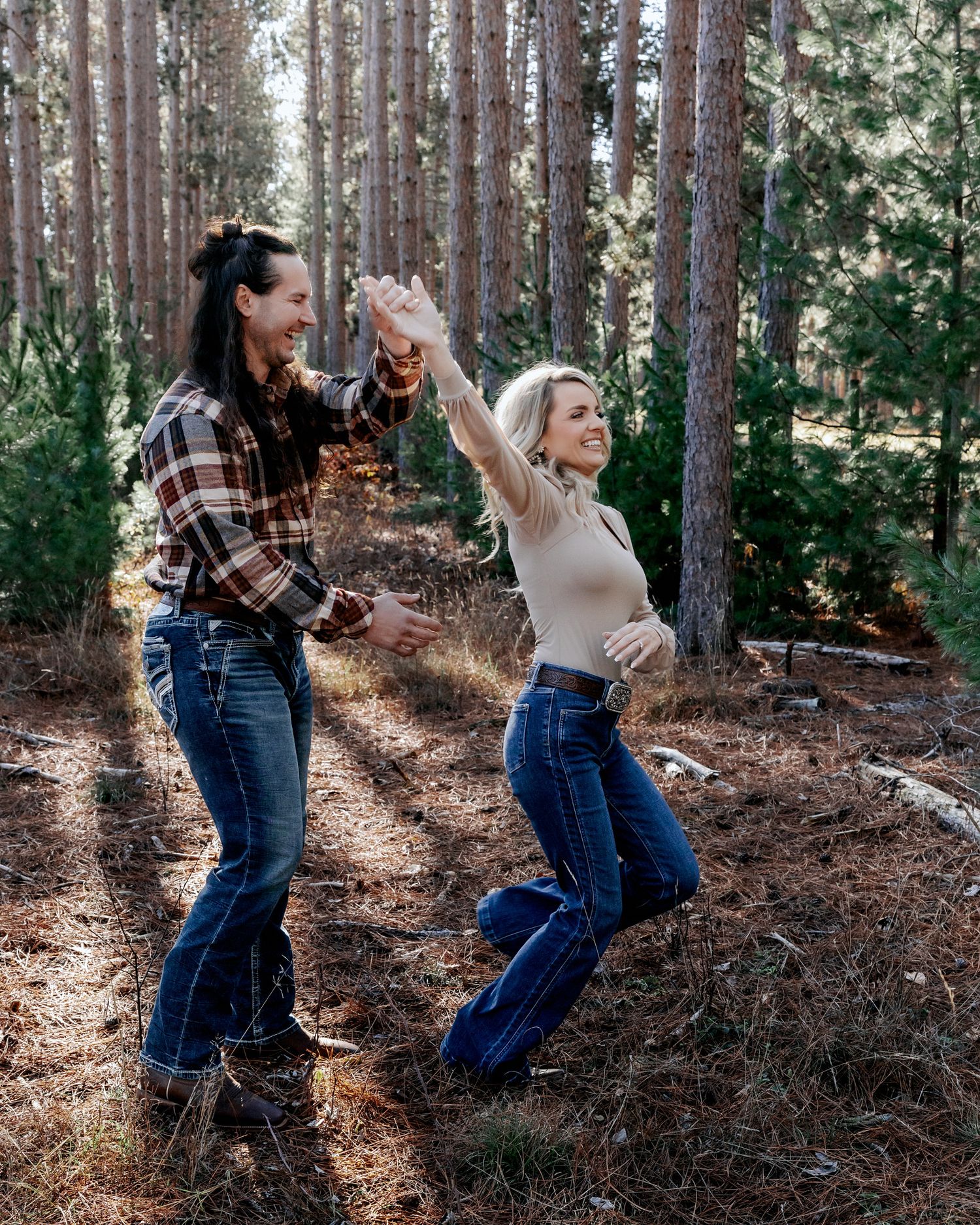 Two women dancing joyfully together in a sunlit pine forest, wearing jeans and casual tops.