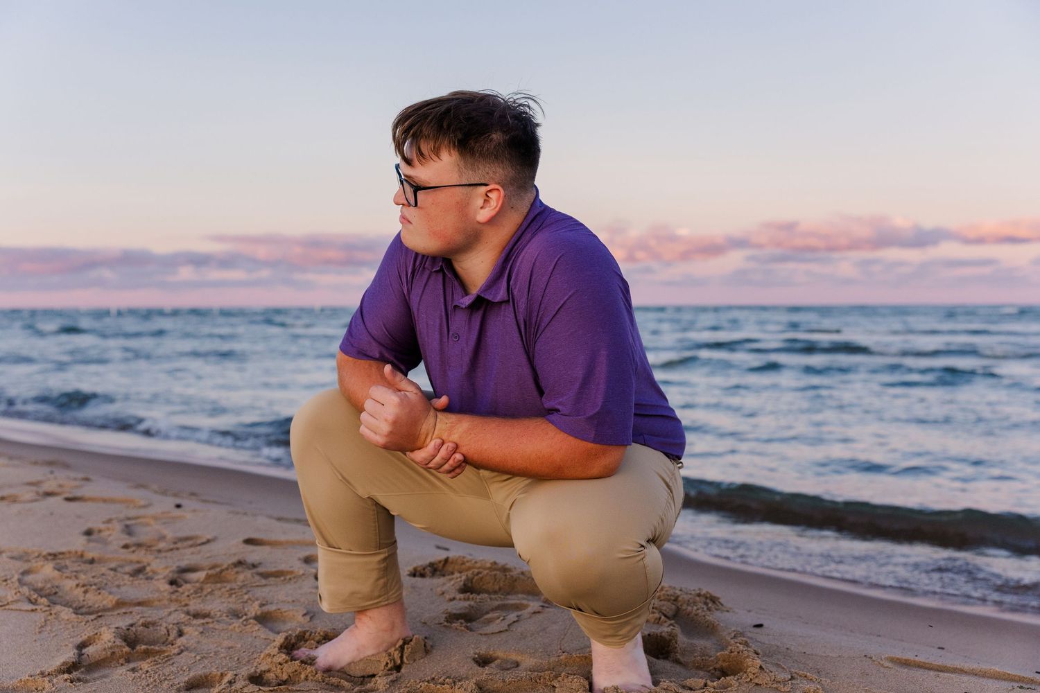 A young man in a purple polo shirt squats on a sandy beach at sunset, gazing toward the ocean waves.